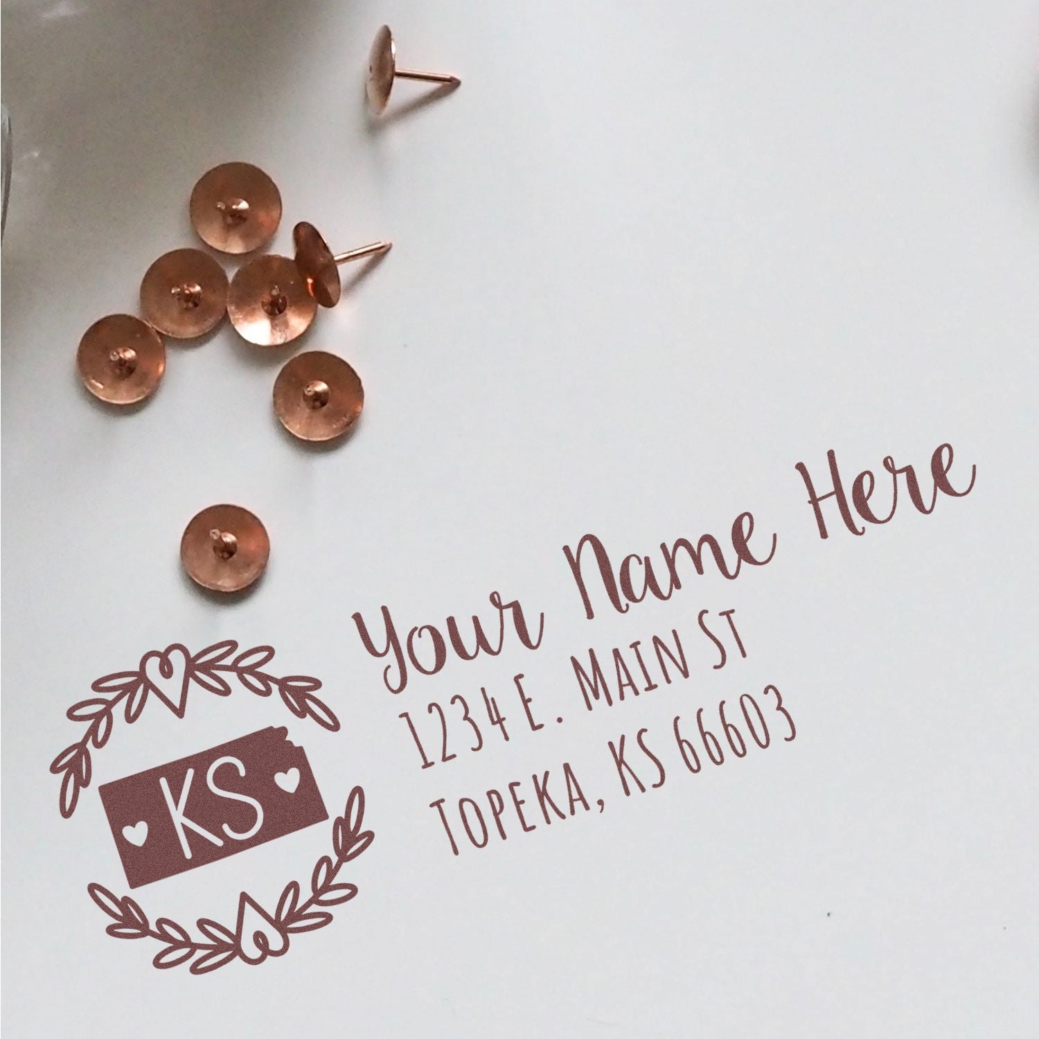 Kansas State Custom Return Address Stamp on white paper with decorative border, featuring placeholder text Your Name Here and an address in Topeka, KS. Copper thumbtacks are scattered nearby.