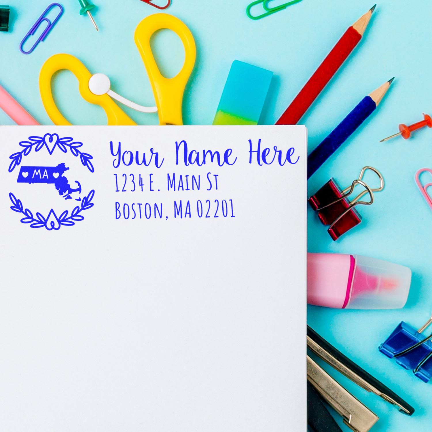 A PSI Pre-Inked Massachusetts State Customized Address Stamp on an envelope, surrounded by colorful office supplies like scissors, pencils, and paper clips on a blue background.