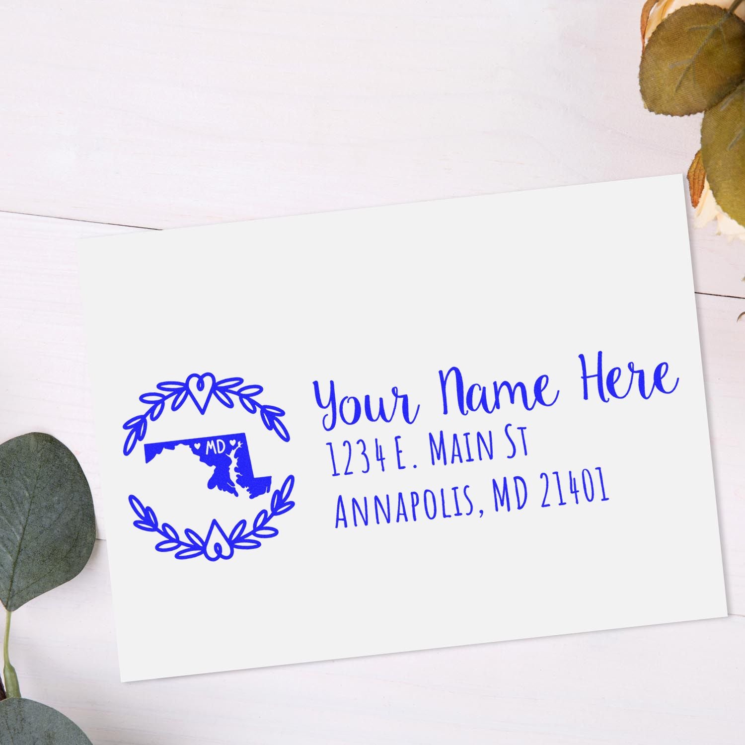 Maryland State Custom Return Address Stamp on white envelope with blue ink, featuring a map outline and decorative border, placed on a light wooden surface with leaves nearby.