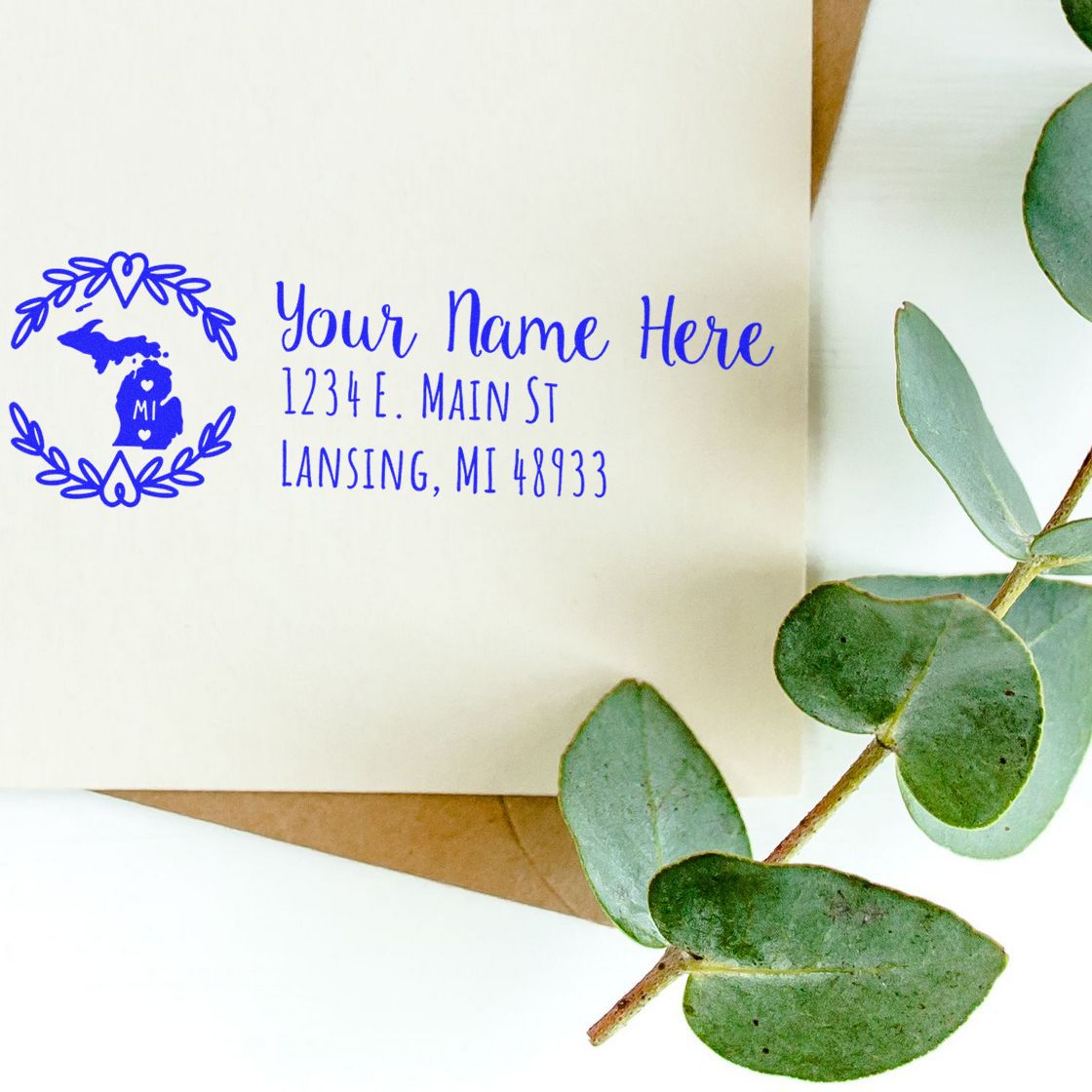 PSI Pre-Inked Michigan State Customized Address Stamp on an envelope, featuring blue text and a Michigan state outline, next to eucalyptus leaves.