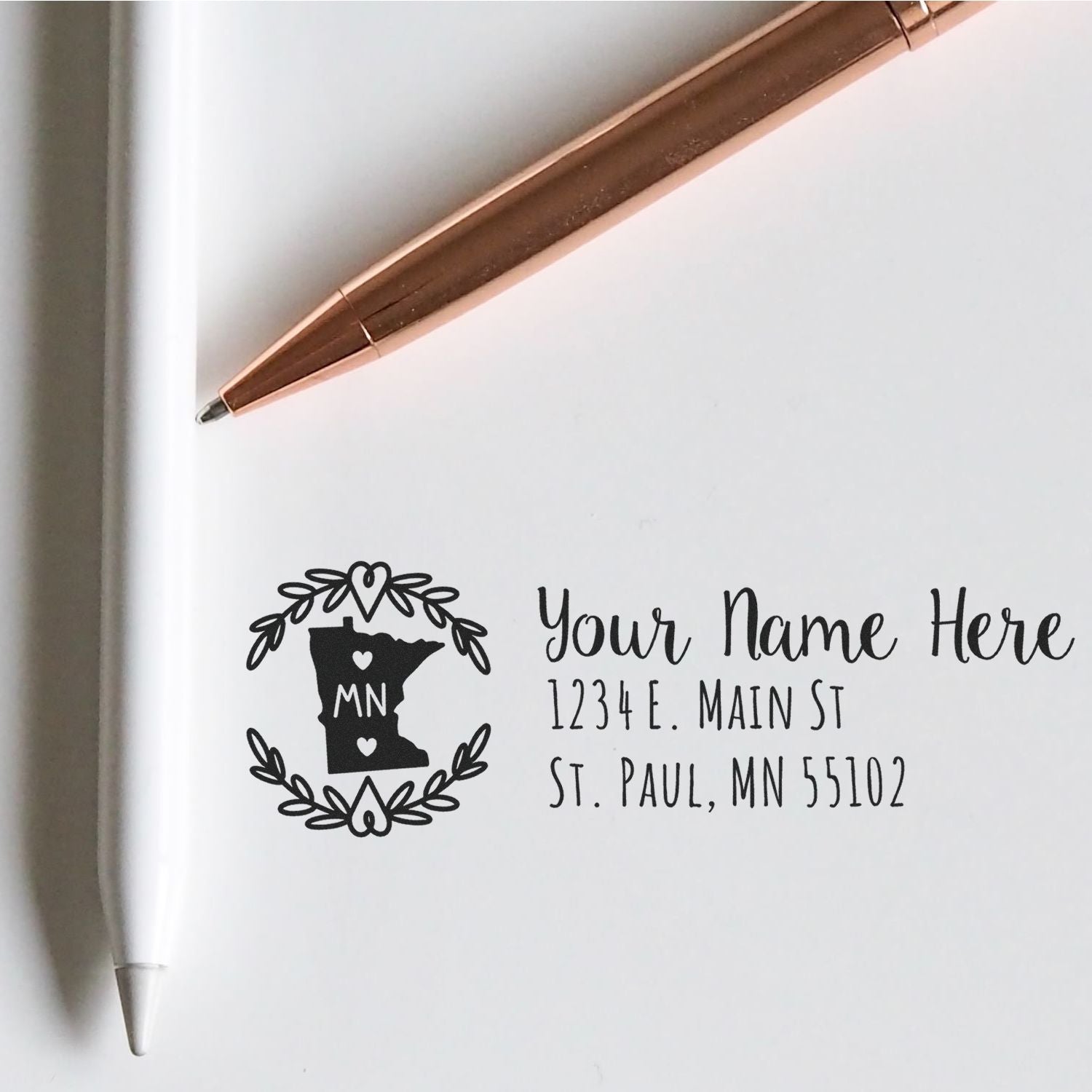 Self-Inking State Wreath of Minnesota Address Stamp on white paper, next to a rose gold pen. The stamp features a Minnesota outline with a wreath, customizable name, and address fields.