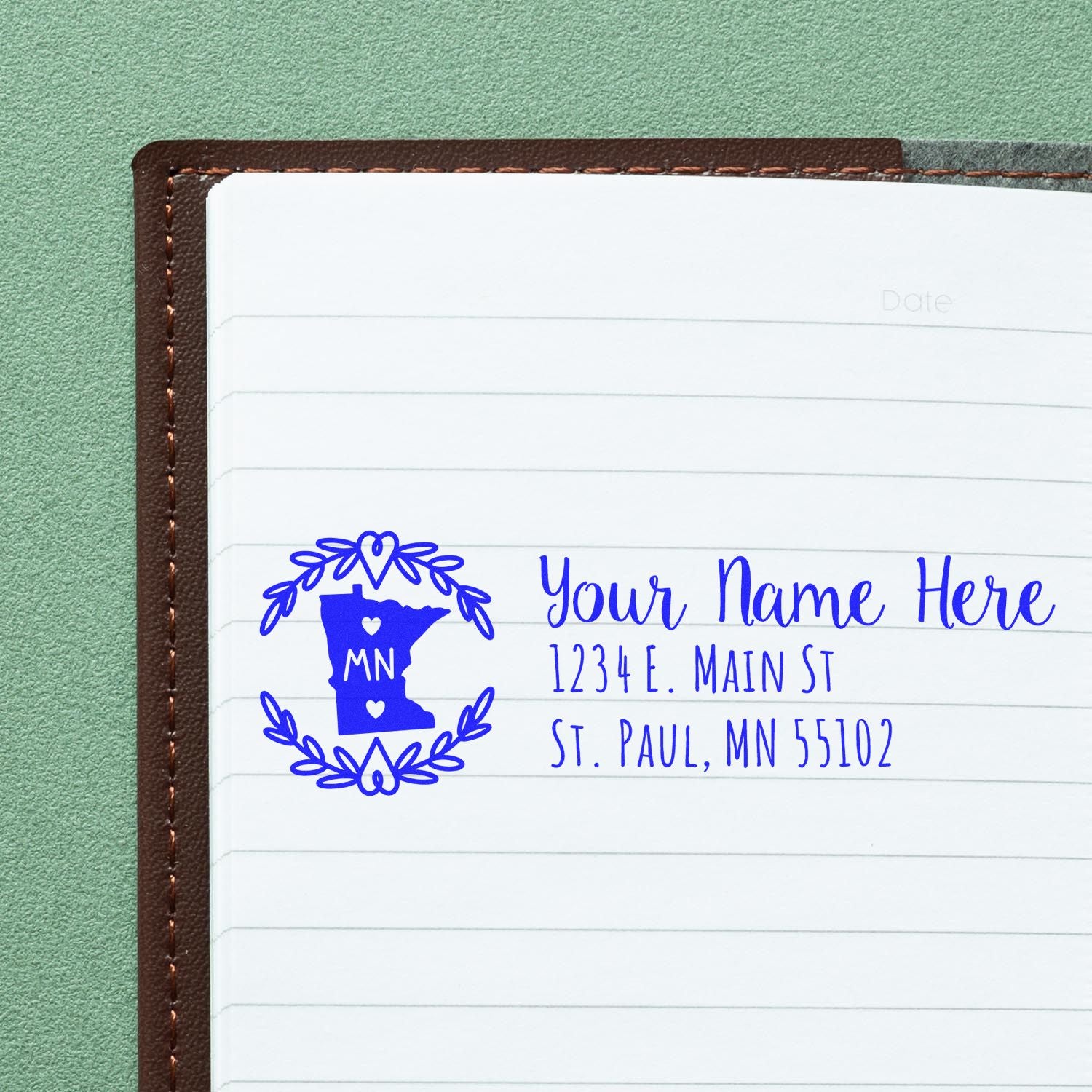 Minnesota State Custom Return Address Stamp in blue ink on a notebook page, featuring a heart and laurel design with sample text: Your Name Here, 1234 E. Main St, St. Paul, MN 55102.