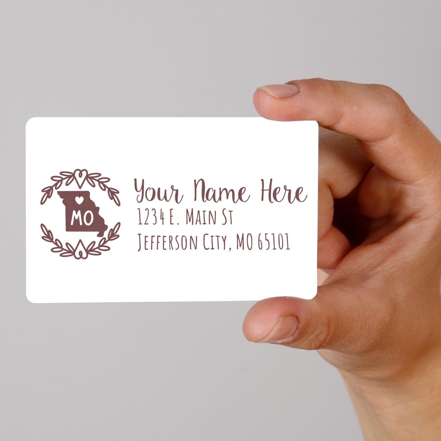 Hand holding a card with a Missouri State Custom Return Address Stamp design, featuring a heart and MO inside a state outline, with sample address text in elegant font.