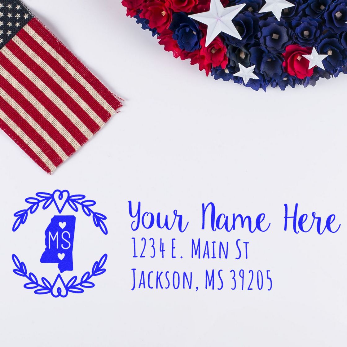 PSI Pre-Inked Mississippi State Customized Address Stamp on white paper with blue text, featuring a heart design and state outline. Nearby are a fabric American flag and a patriotic floral wreath.