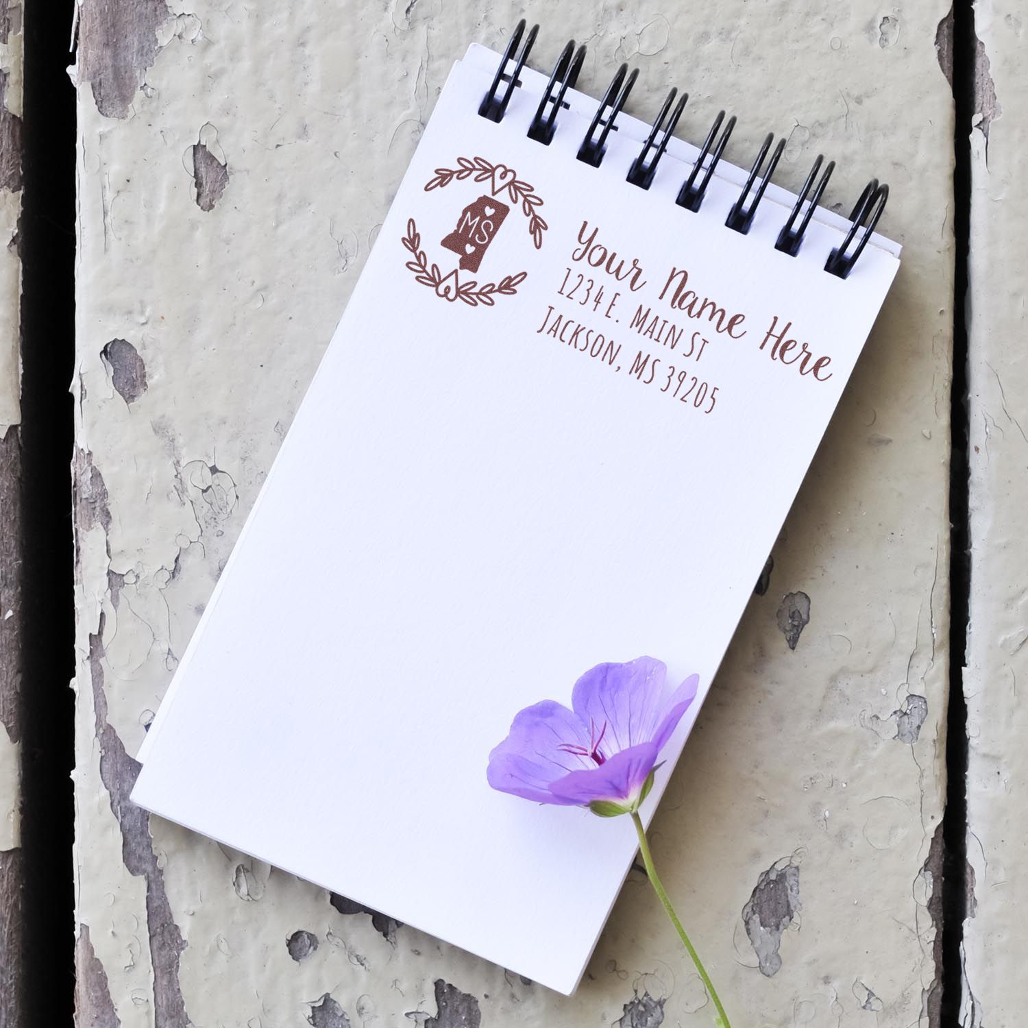 Notepad with a stamped address using the Mississippi State Custom Return Address Stamp, featuring a floral design and placeholder text. A purple flower rests on the notepad, placed on a rustic surface.