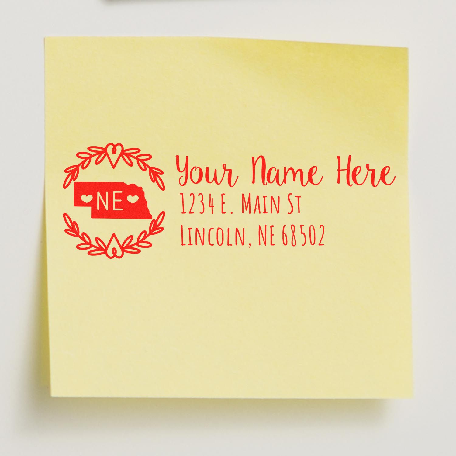 Self-Inking State Wreath of Nebraska Address Stamp on a yellow sticky note, featuring a red wreath design with NE and customizable address lines in elegant font.