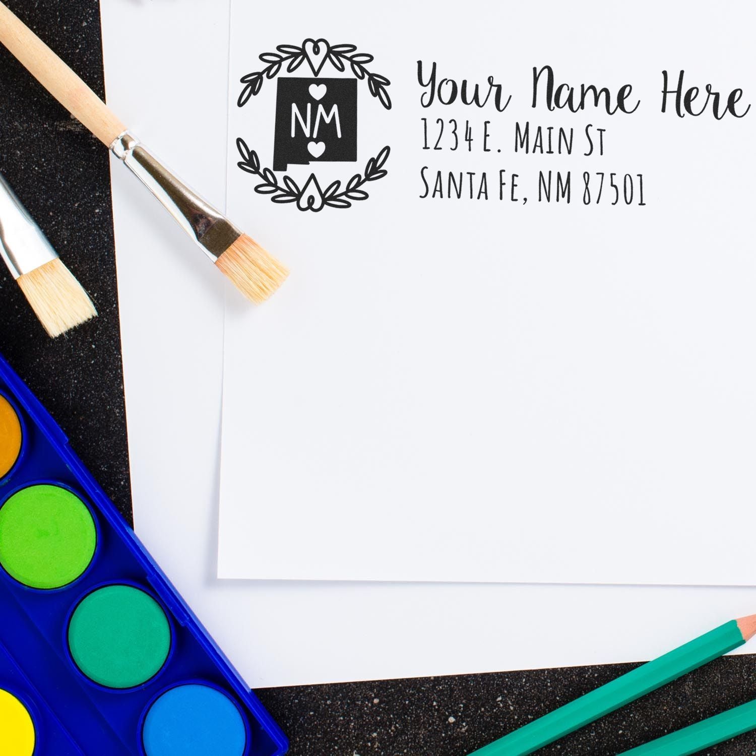 PSI Pre-Inked New Mexico State Customized Address Stamp on white paper with paintbrushes and watercolor set, showcasing a personalized address in elegant font and decorative border.