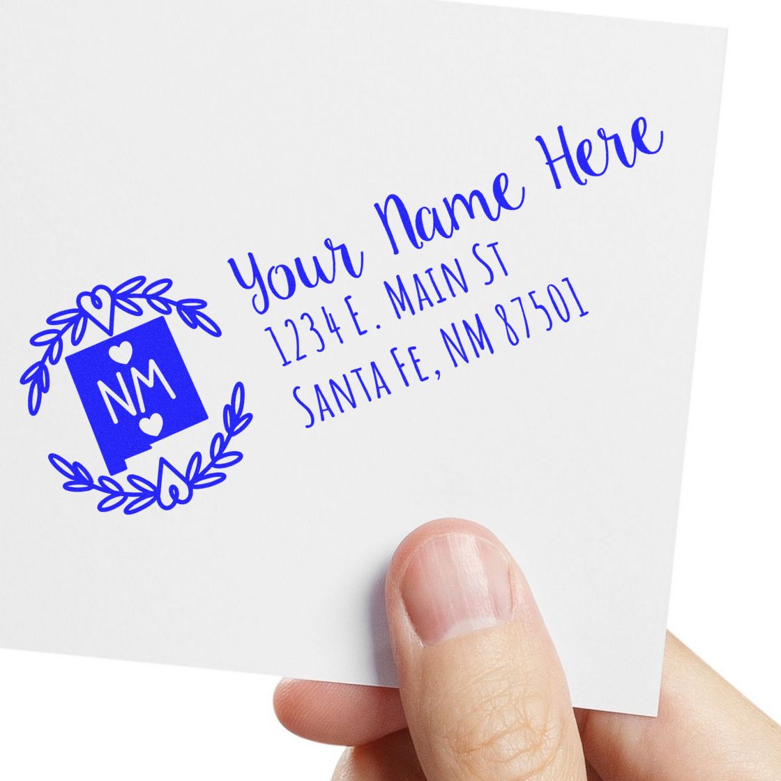 A hand holds an envelope featuring the New Mexico State Custom Return Address Stamp in blue, with a decorative NM emblem and sample address text in elegant script.