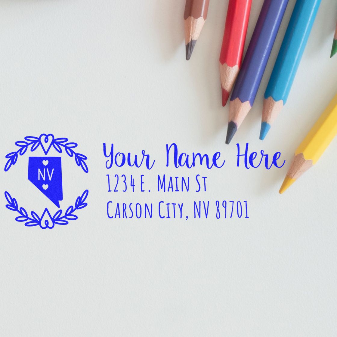 Nevada State Custom Return Address Stamp on white paper with colorful pencils. Features a blue Nevada state outline and sample address text in elegant font. Perfect for personalized mailings.