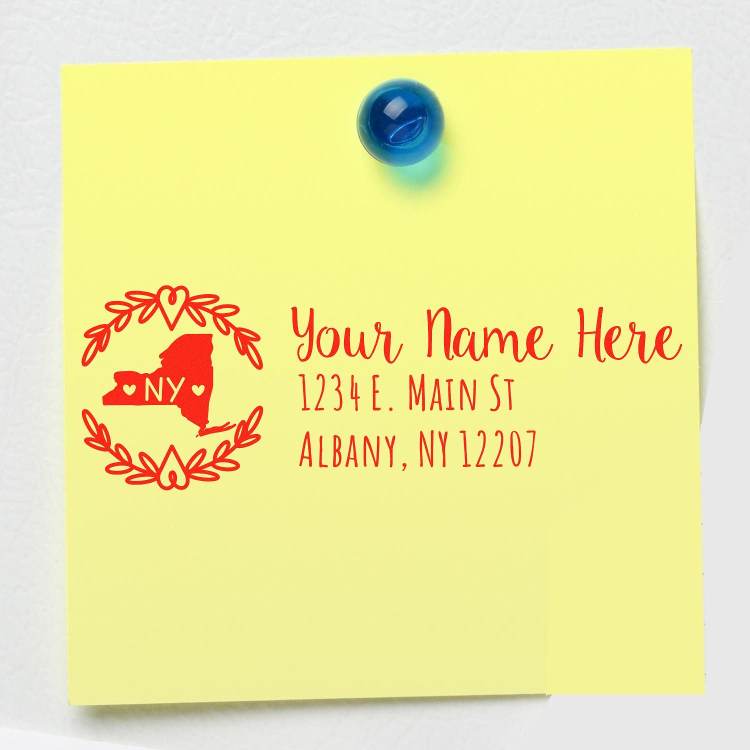 Yellow note with red text showing a sample address using the PSI Pre-Inked New York State Customized Address Stamp, featuring a NY state outline and decorative border, pinned with a blue magnet.