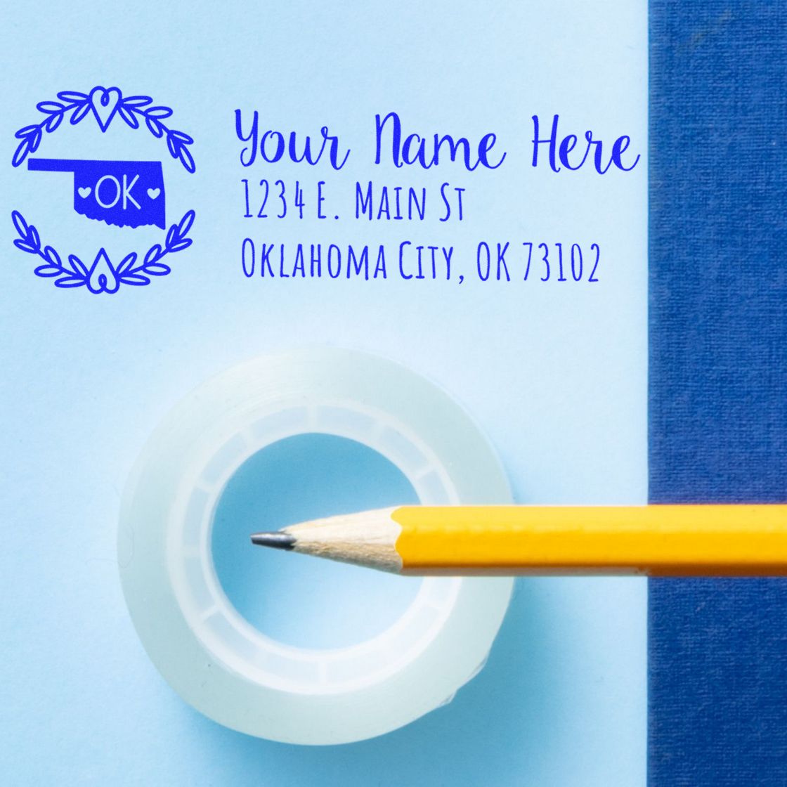 Self-Inking State Wreath of Oklahoma Address Stamp on blue paper with a pencil and tape, displaying a sample address in blue ink. Perfect for personalized mailings.