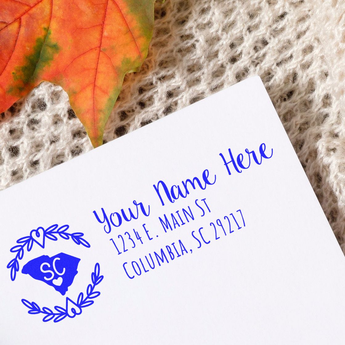 South Carolina State Custom Return Address Stamp on white envelope with blue ink, featuring a heart and state outline. Placed on a textured surface with an autumn leaf nearby.