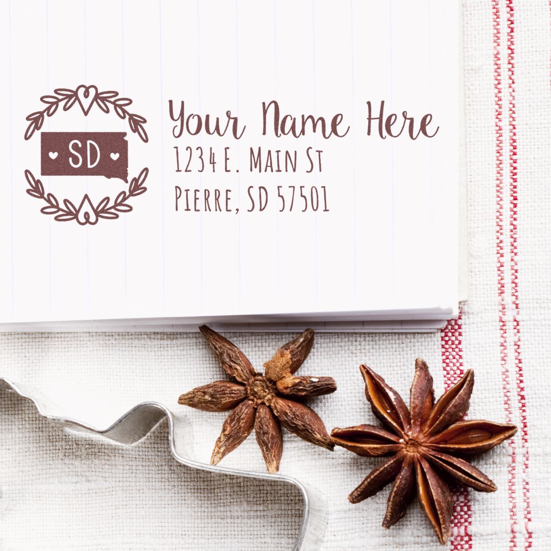 Slim South Dakota Personalized Pre-Inked Address Stamp on a notepad with a decorative border, featuring sample text. Surrounded by star anise and a cookie cutter on a textured surface.