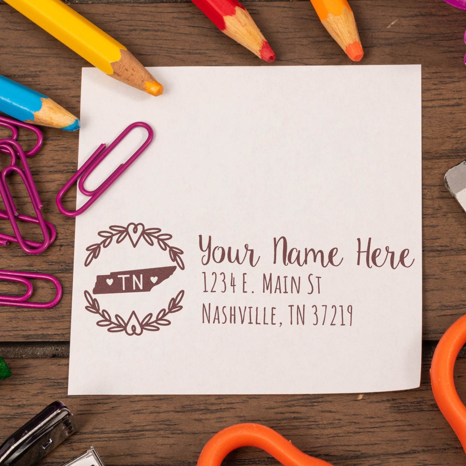 Tennessee State Custom Return Address Stamp on a white paper with decorative border, surrounded by colorful pencils, paperclips, and a wooden desk background.