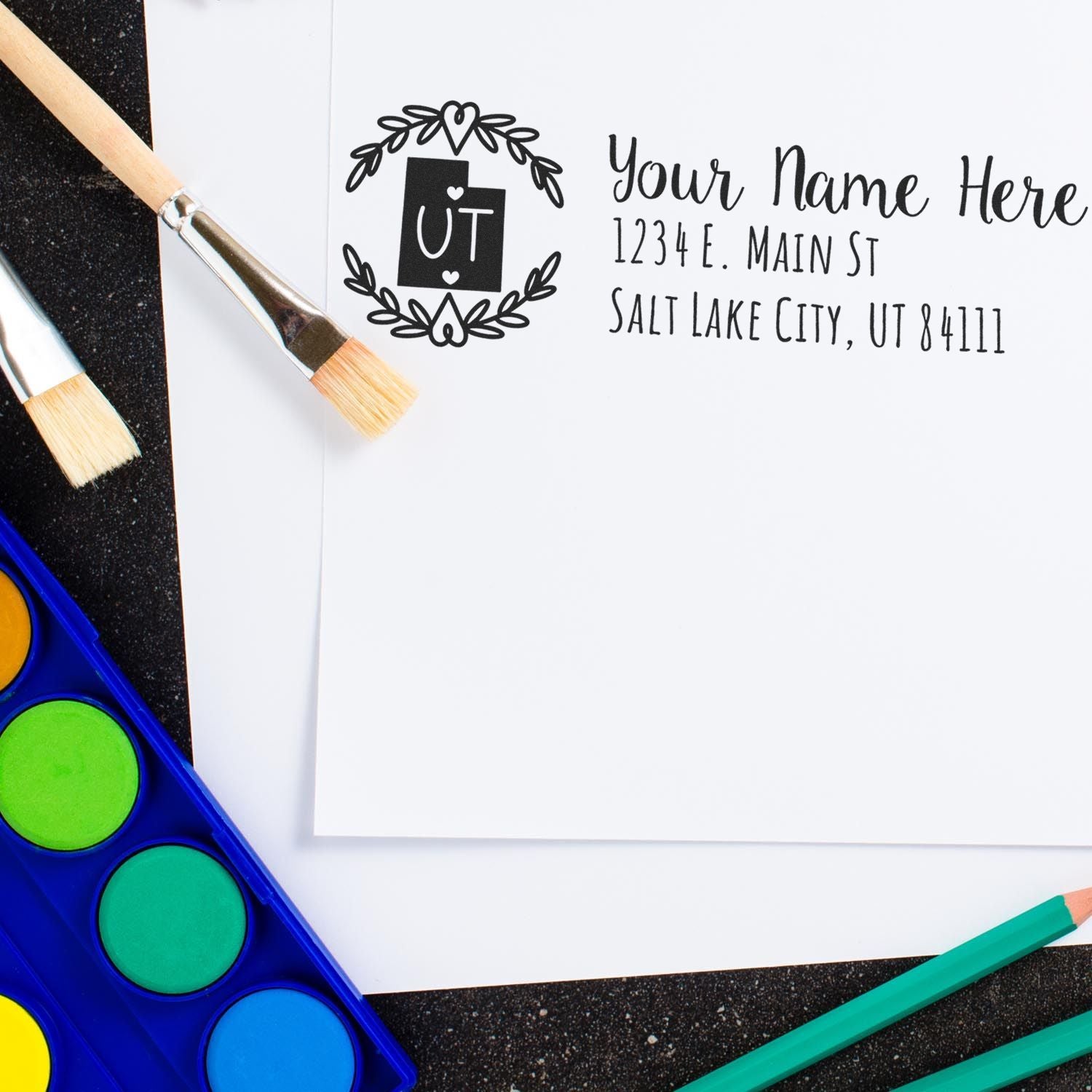 Utah State Custom Return Address Stamp on white paper with a heart and UT design, surrounded by paintbrushes and watercolor paints.