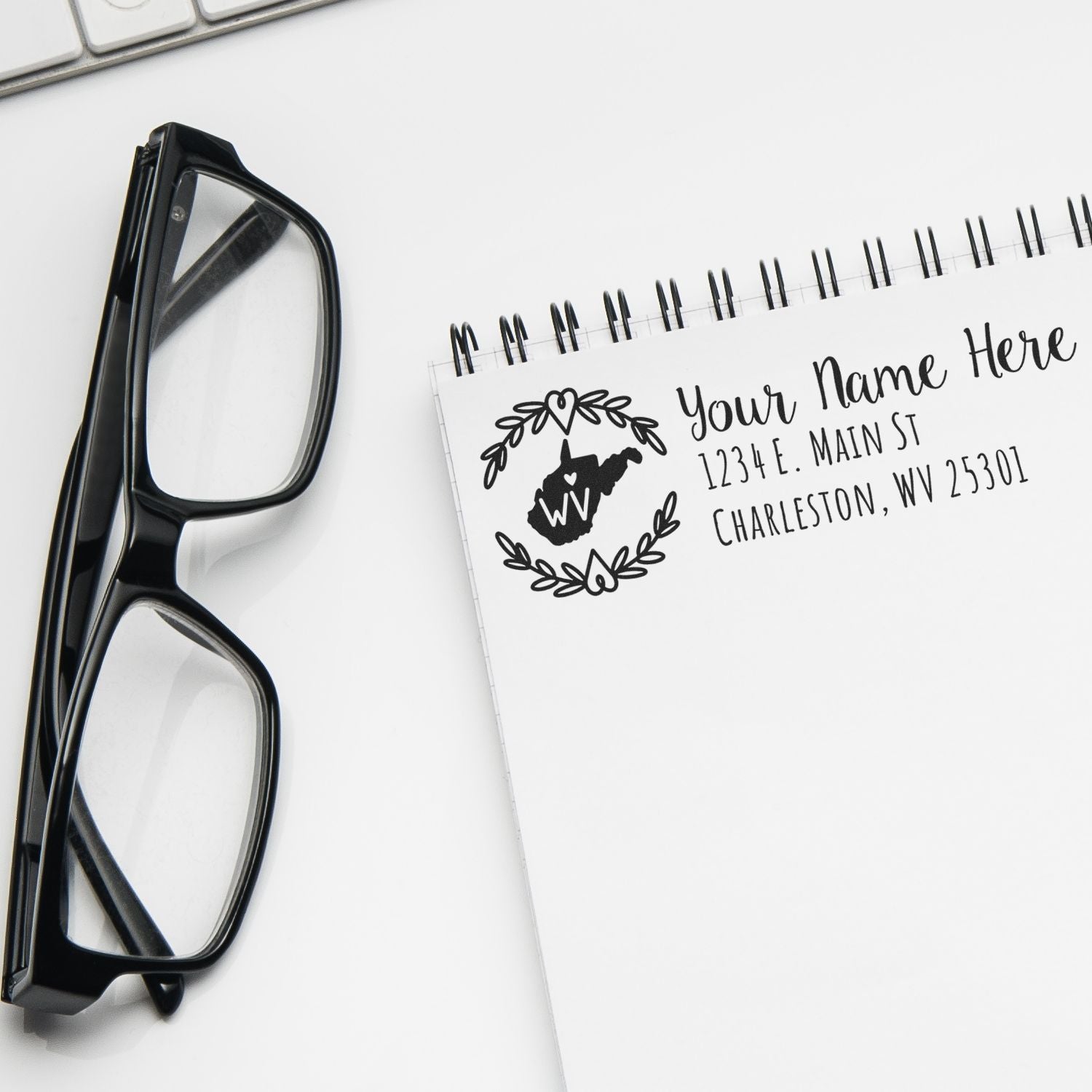 Self-Inking State Wreath of West Virginia Address Stamp on a notepad, next to black glasses and a keyboard, displaying a sample address with a decorative state outline and wreath design.