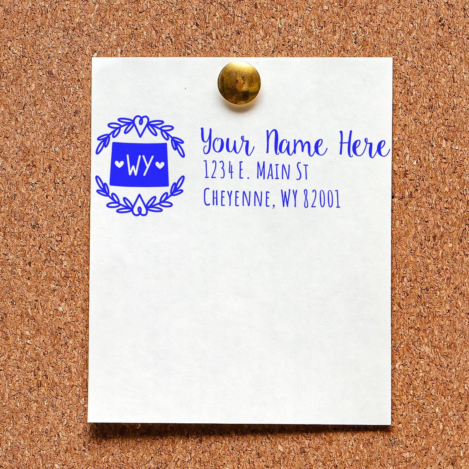 Self-Inking State Wreath of Wyoming Address Stamp on a corkboard, displaying a sample address with a decorative wreath design and WY initials in blue ink.