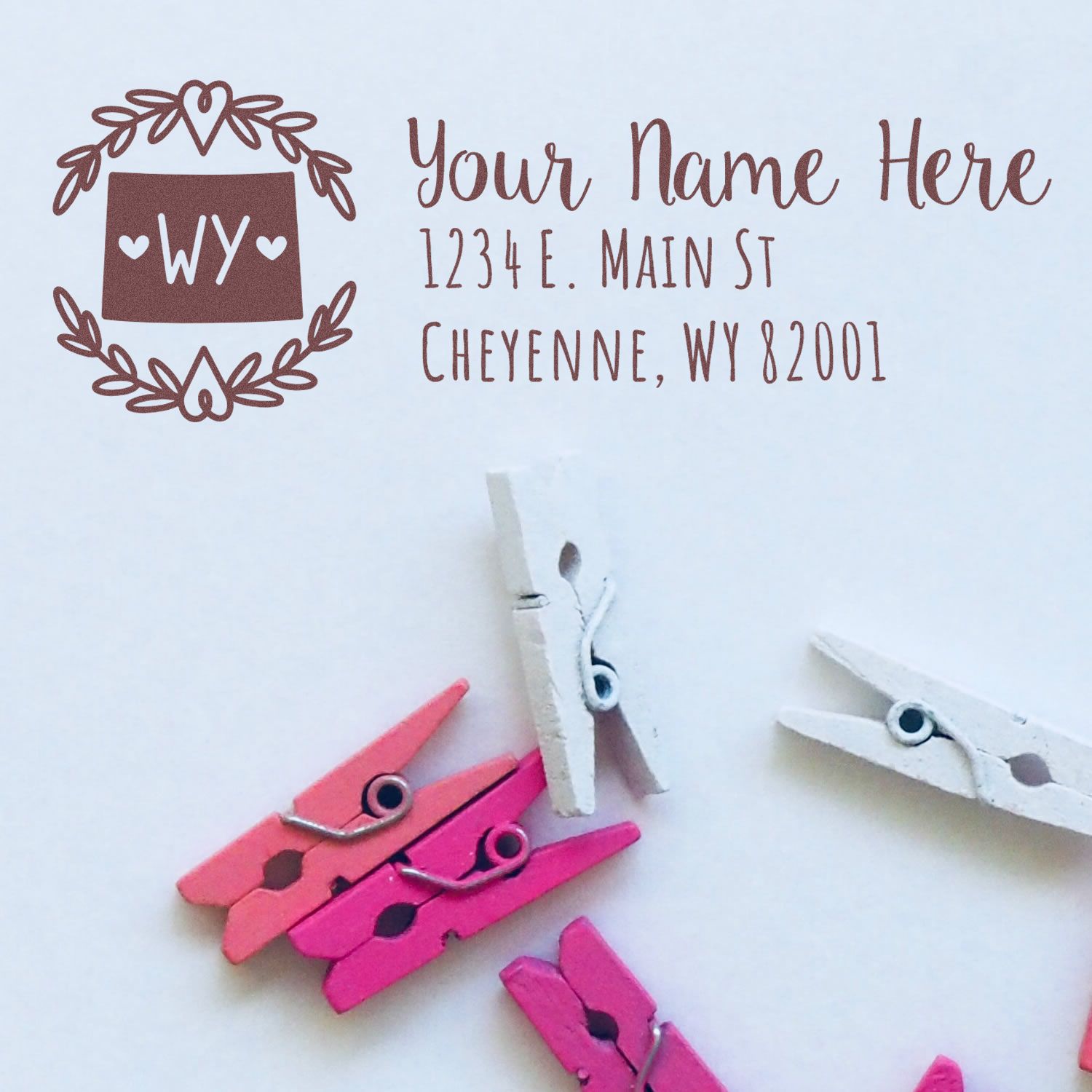 Wyoming State Custom Return Address Stamp with a decorative border, featuring sample text for a Cheyenne address. Pink and white clothespins are scattered below the stamp design.