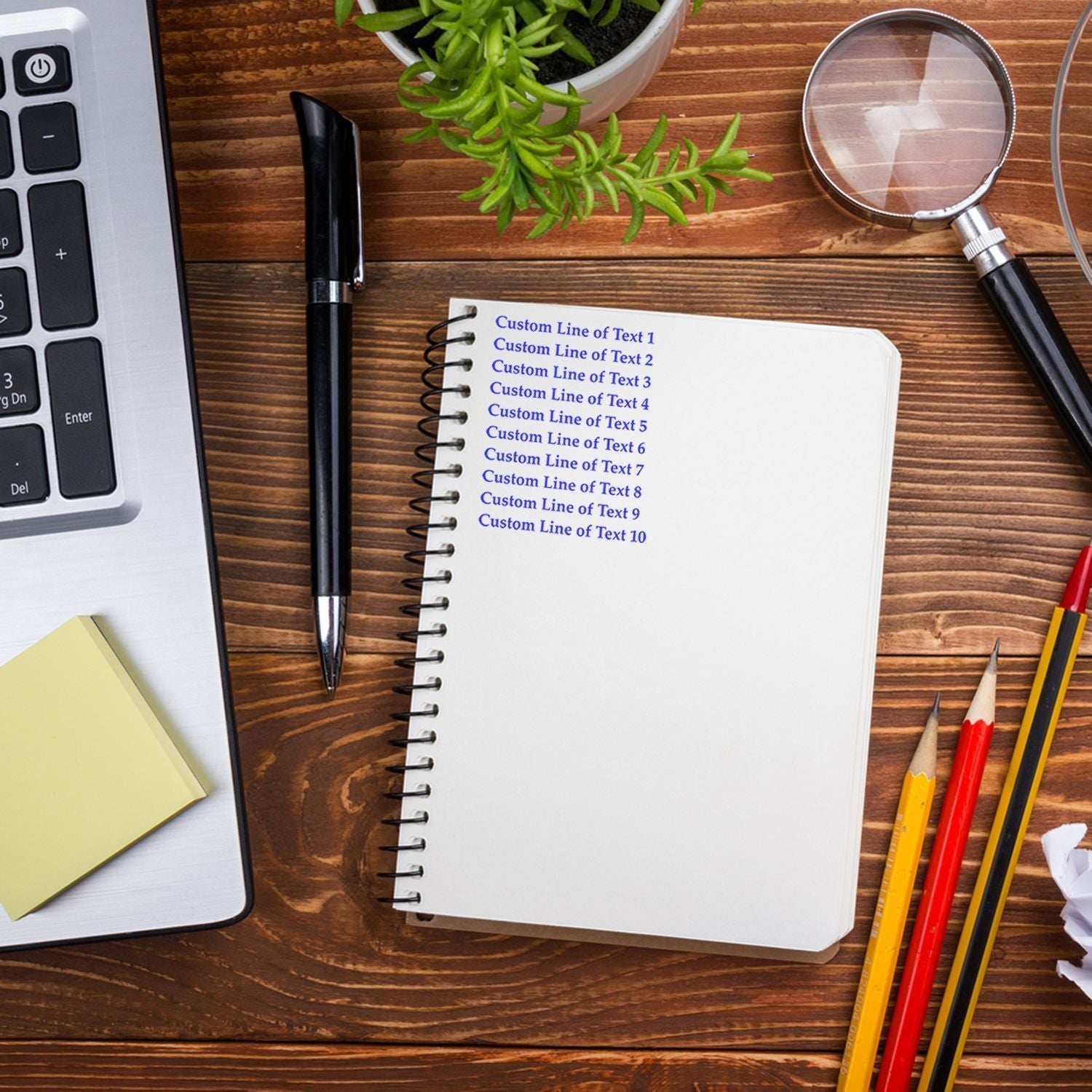 10 Line Custom Rubber Stamp with Wood Handle used on a notebook, surrounded by a laptop, pen, magnifying glass, plant, and pencils.