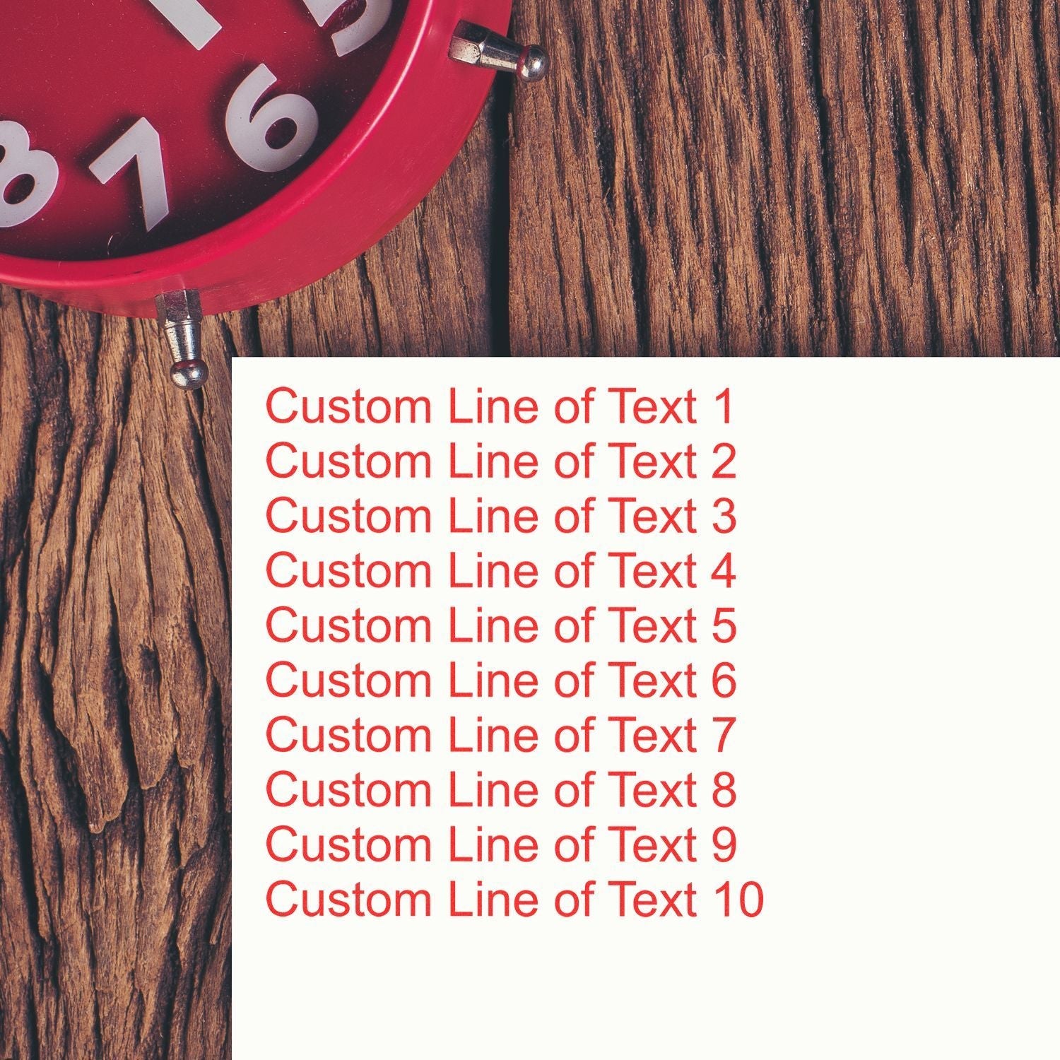 10 Line Custom Rubber Stamp with Wood Handle in use, stamping red text on white paper, placed on a wooden surface next to a red clock.