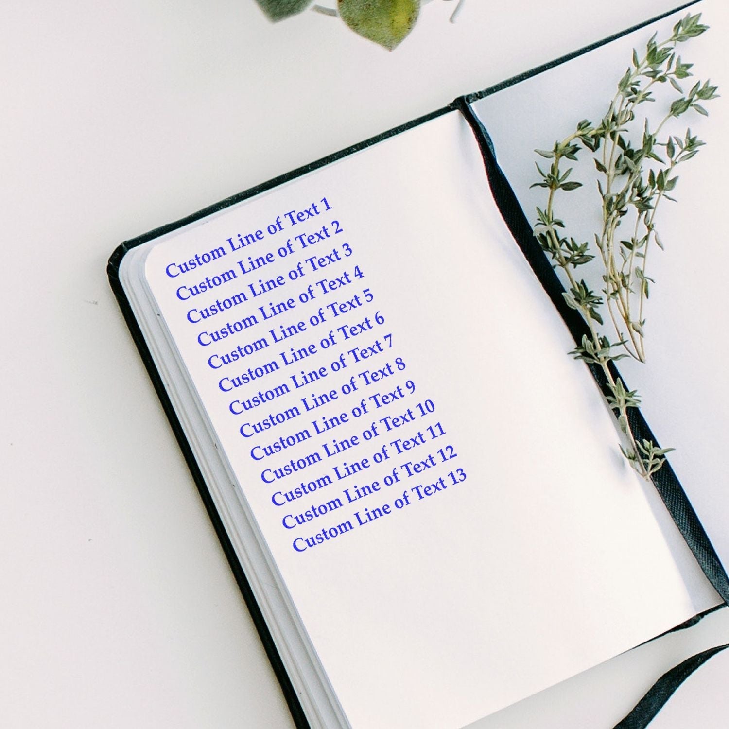 13 Line Custom Rubber Stamp with Wood Handle used on a notebook, displaying 13 lines of blue text. A sprig of greenery lies beside the notebook.