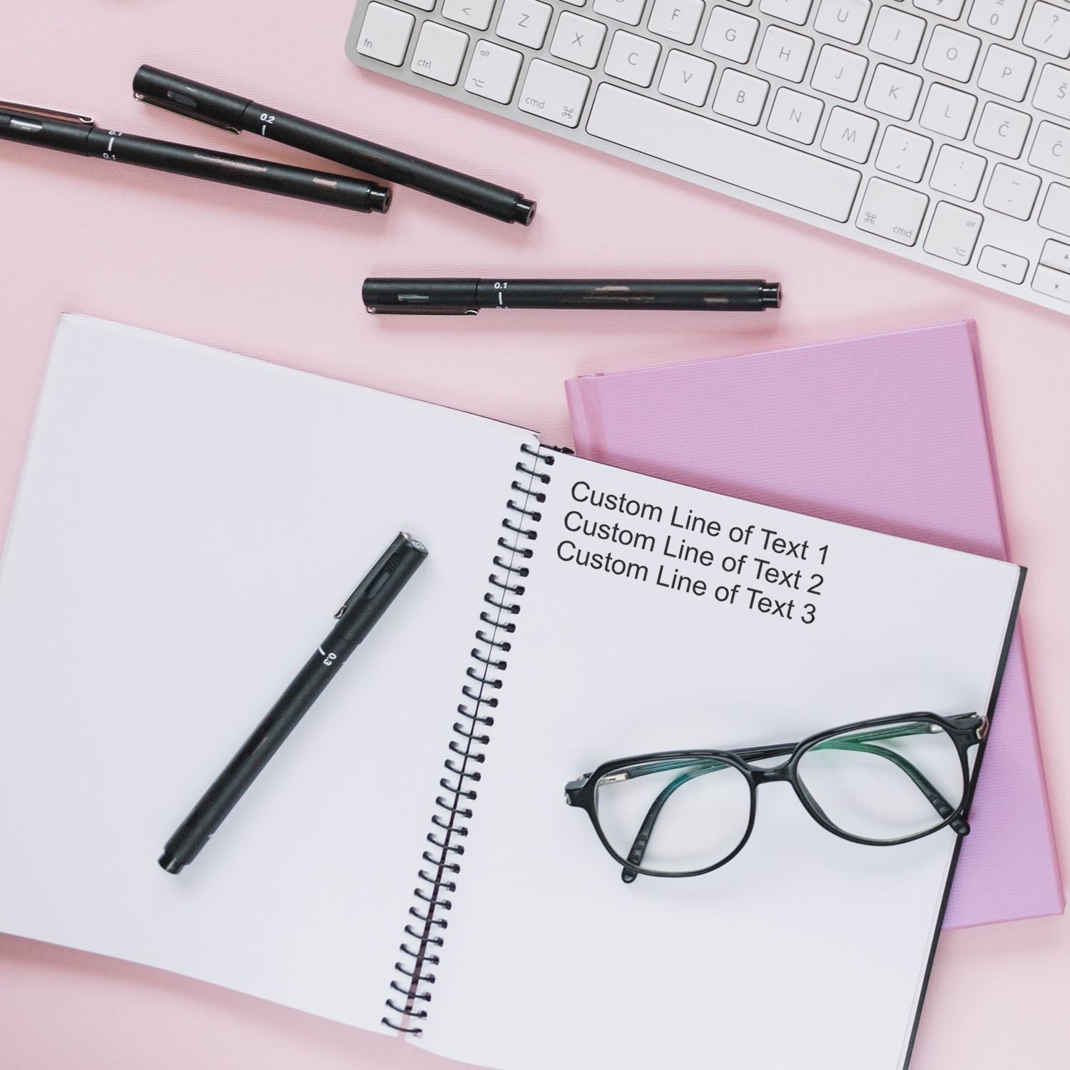 3 Line Custom Rubber Stamp with Wood Handle used on a notebook, surrounded by pens, glasses, and a keyboard on a pink surface.