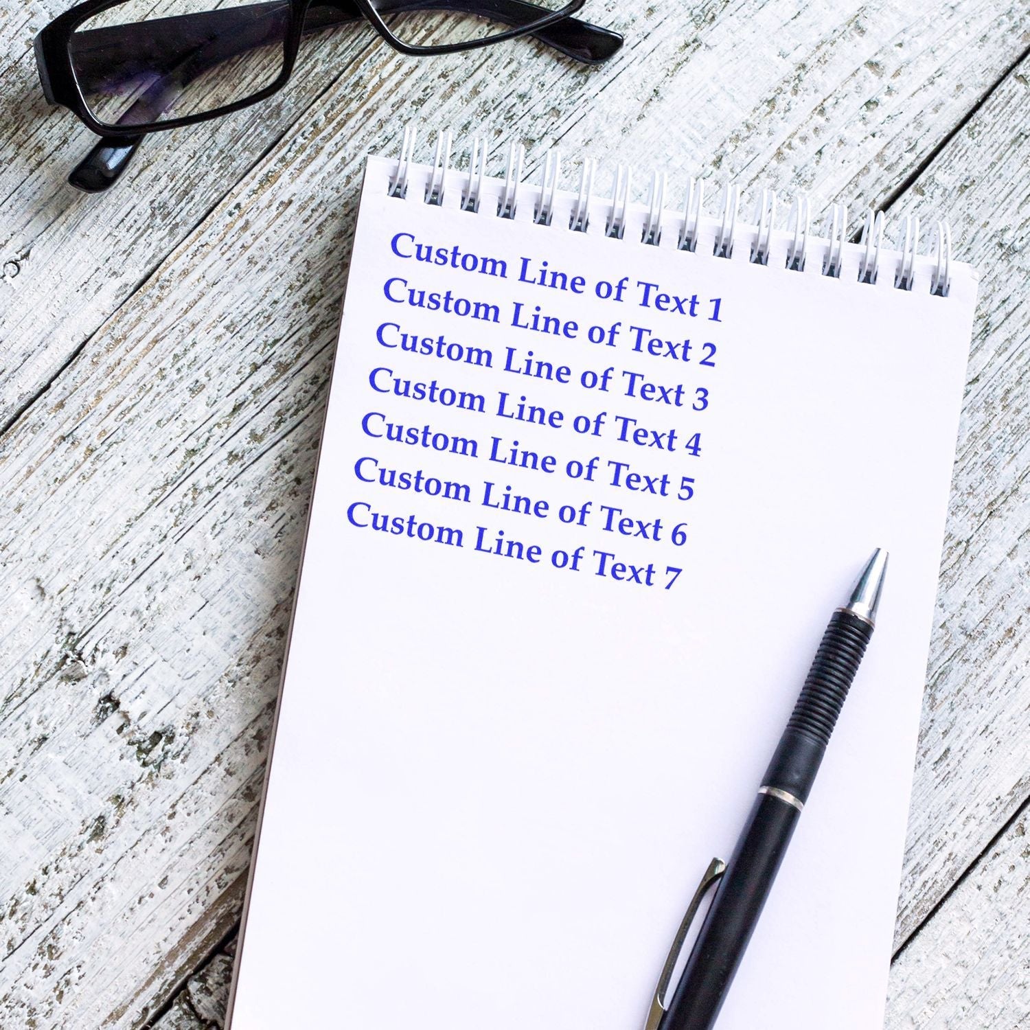 A 7 Line Custom Rubber Stamp with Wood Handle is used on a notepad with custom text lines. A pen and glasses are placed nearby.