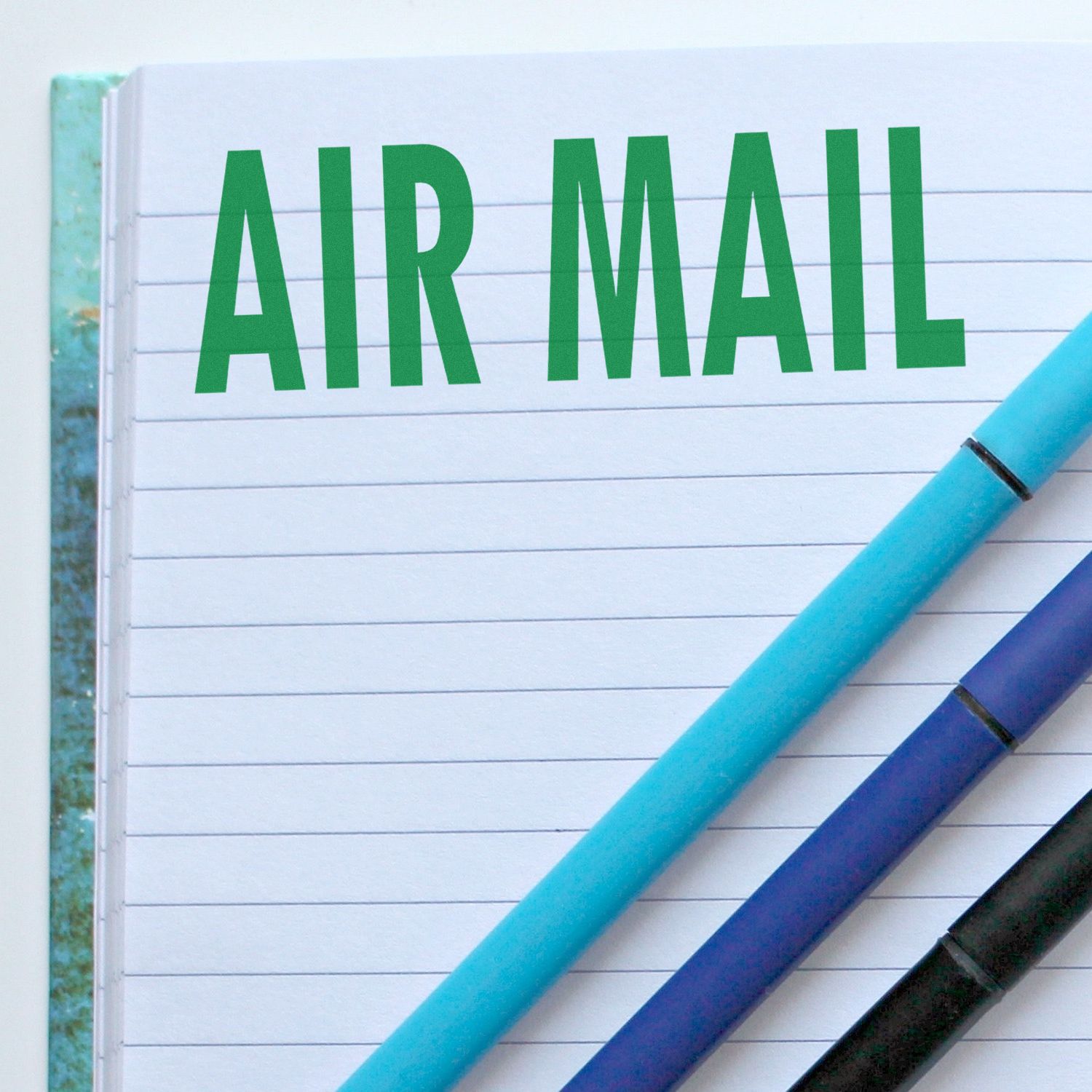 Large Pre-Inked Air Mail Stamp impression on a lined notebook page with three pens placed diagonally across the page.