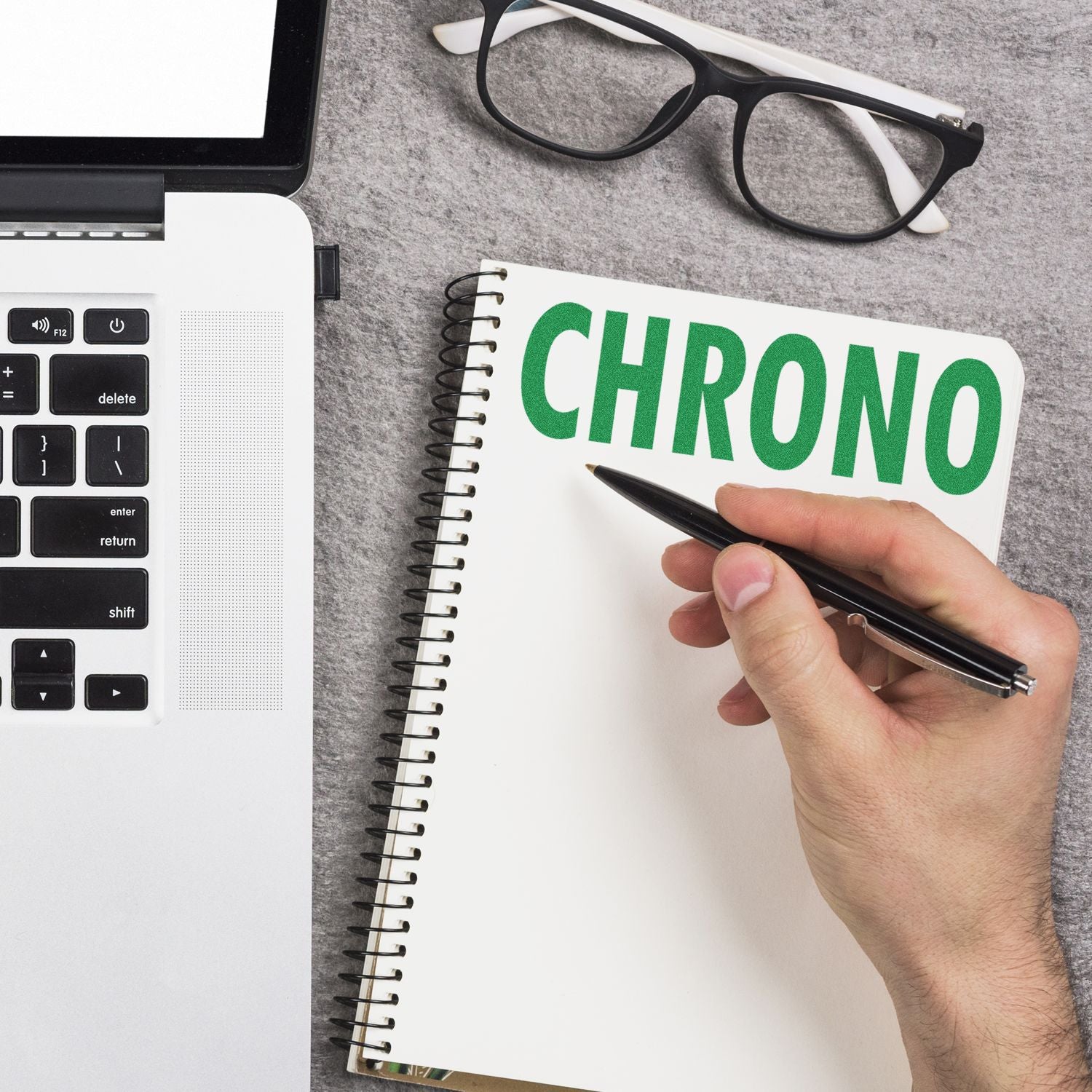 A hand holding a pen near a notebook with CHRONO written on it, beside a laptop and glasses. Large Pre-Inked Chrono Stamp.