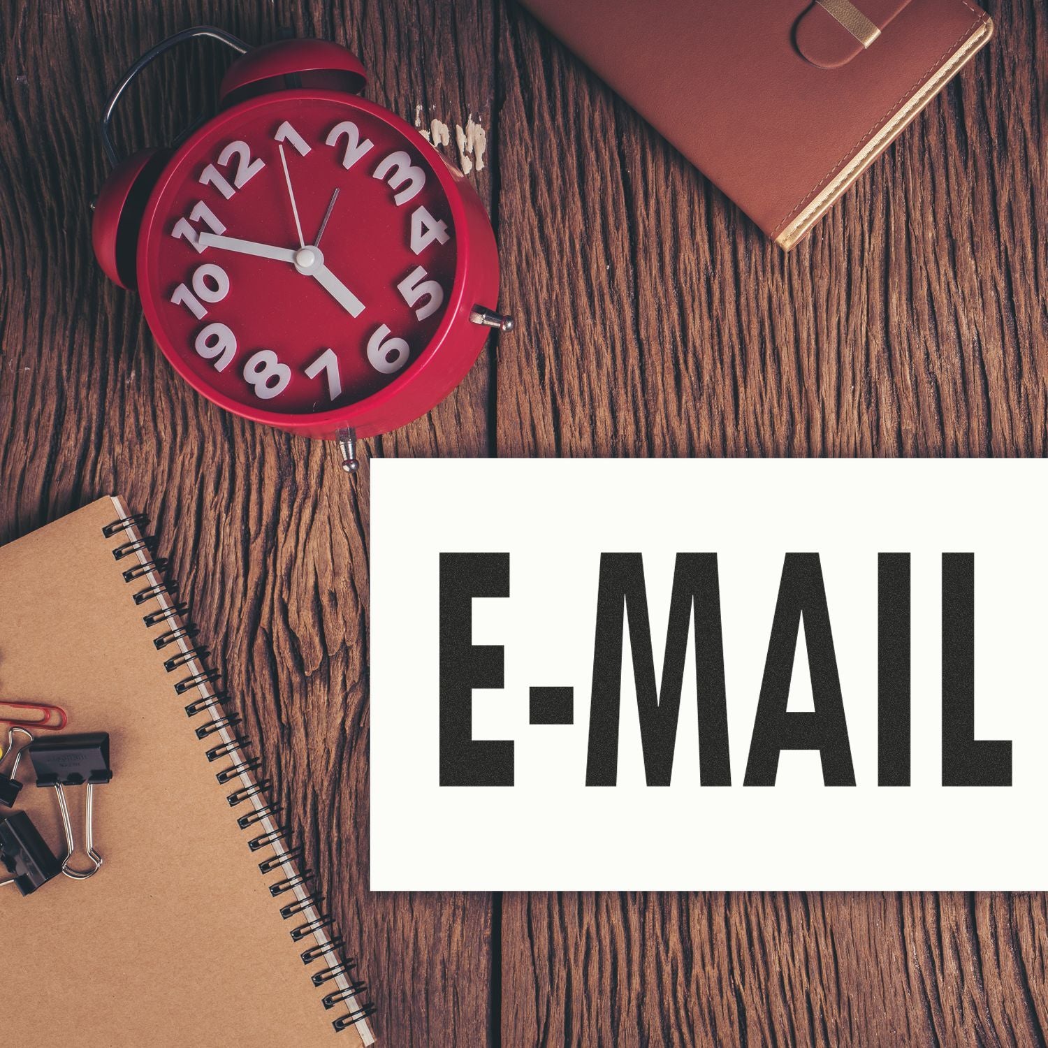 A Large Pre-Inked E Mail Stamp on a wooden desk with a red clock, notebook, and brown leather wallet in the background.