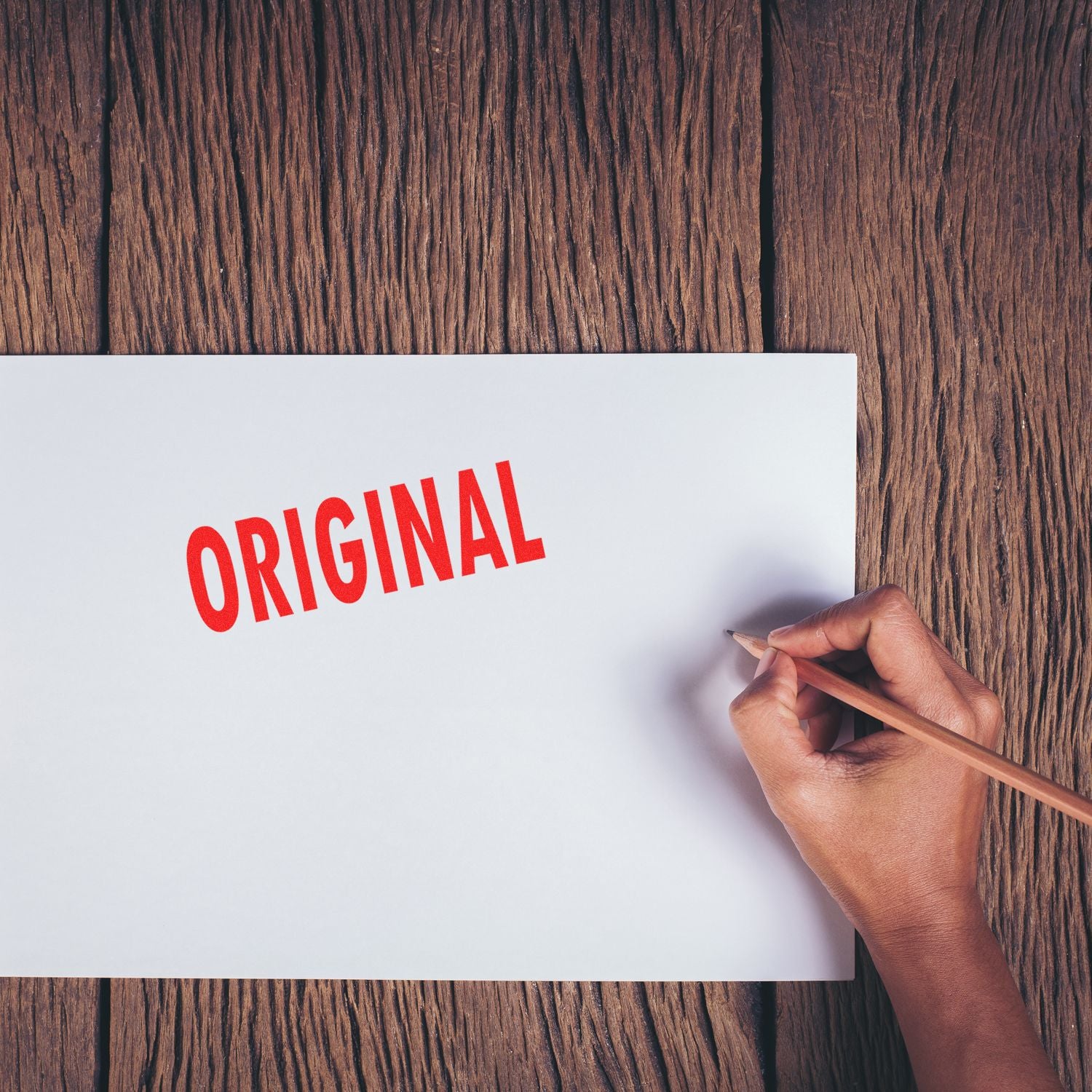 Hand holding a pencil next to a paper stamped with ORIGINAL in red using the Large Pre-Inked Original Stamp on a wooden surface.