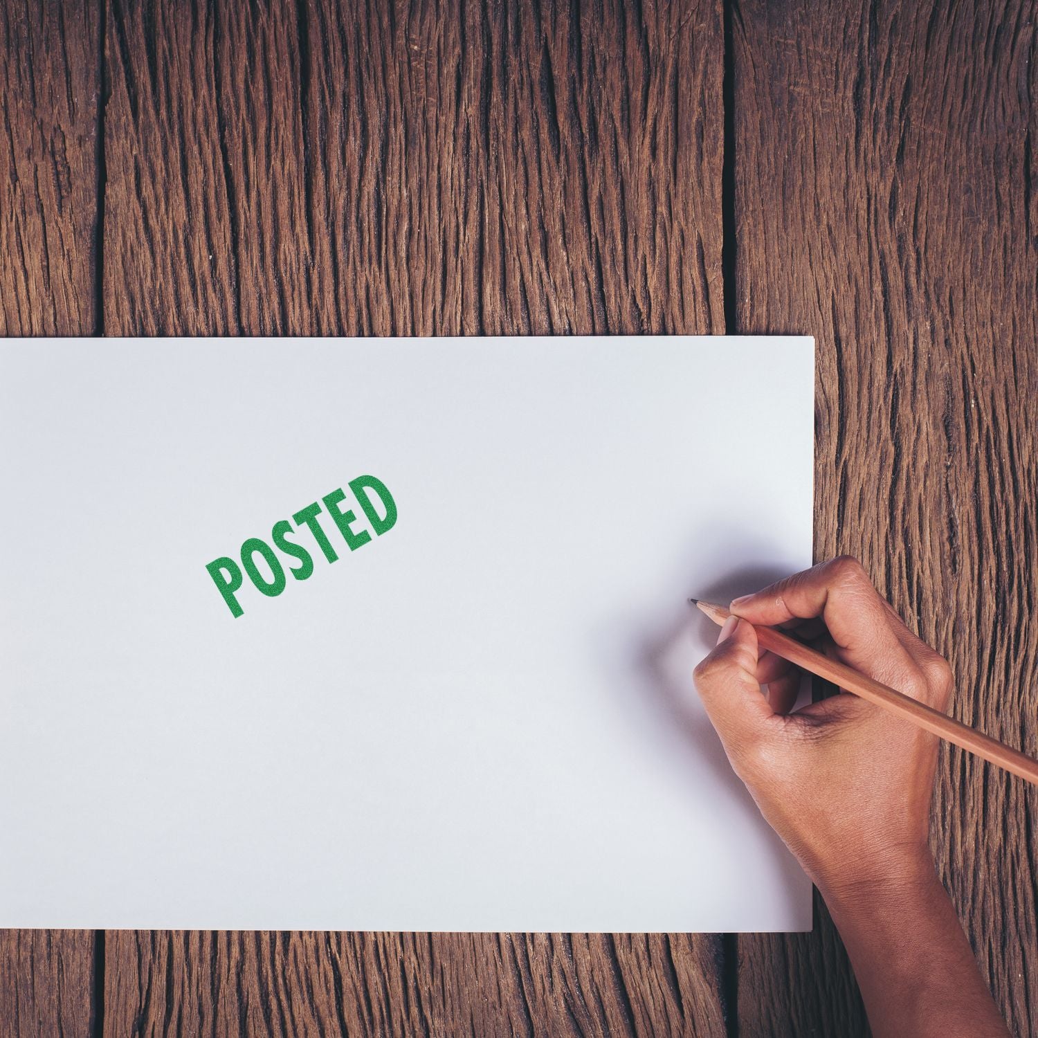 Hand holding a pencil next to a white paper stamped with POSTED in green using a Large Pre-Inked Posted Stamp on a wooden surface.