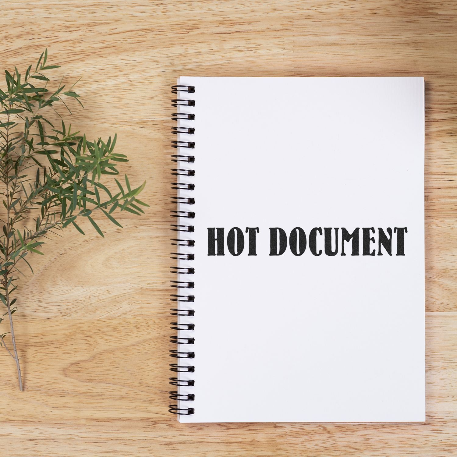 A spiral notebook on a wooden table stamped with HOT DOCUMENT using the Large Pre-Inked Hot Document Stamp, next to a sprig of greenery.