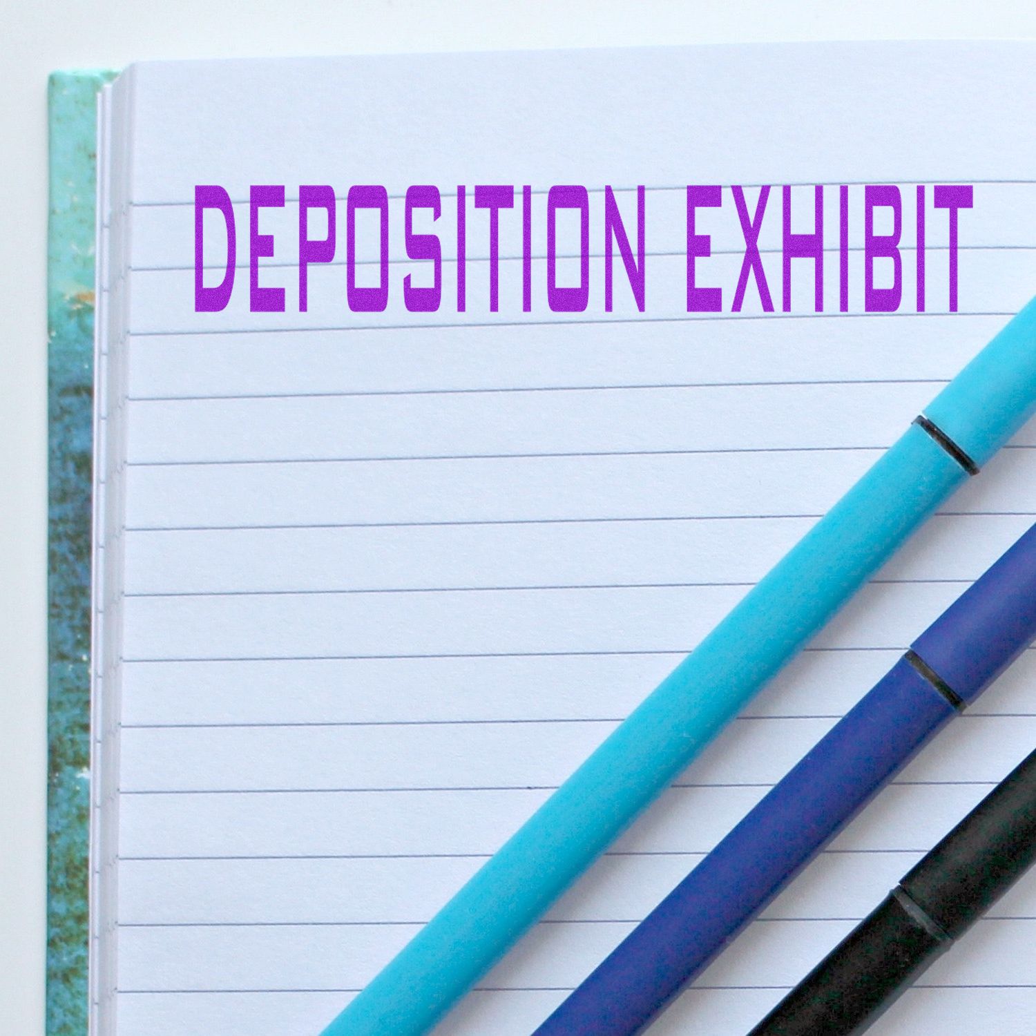 Notebook with DEPOSITION EXHIBIT stamped in purple using the Large Pre-Inked Deposition Exhibit Stamp, with three pens lying on the page.