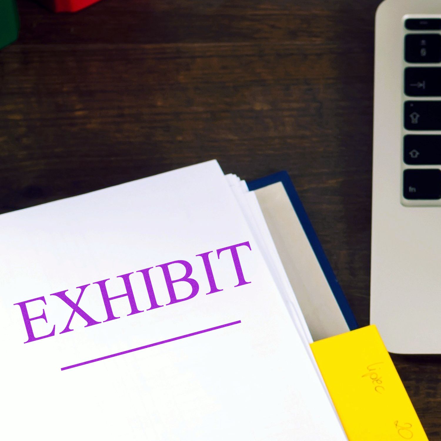 Large Pre-Inked Exhibit Stamp marking a document with EXHIBIT in purple ink, placed on a wooden desk next to a laptop.