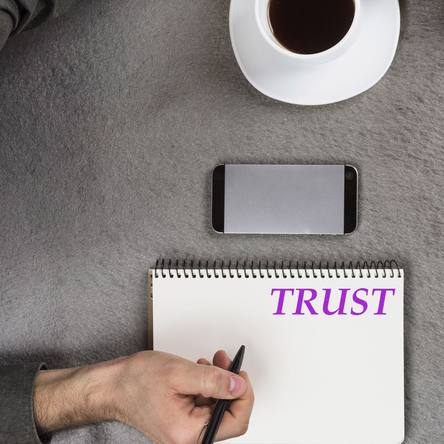 A person writing TRUST in a notebook with a Large Pre-Inked Trust Stamp, next to a smartphone and a cup of coffee on a gray surface.