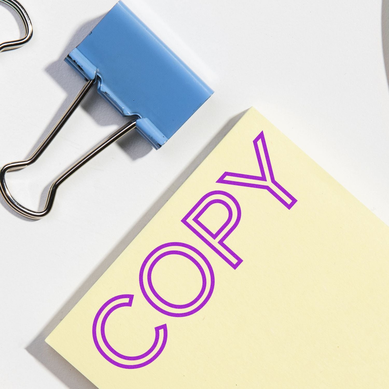 Large Pre-Inked Outline Font Copy Stamp in purple ink on a yellow paper next to a blue binder clip on a white surface.