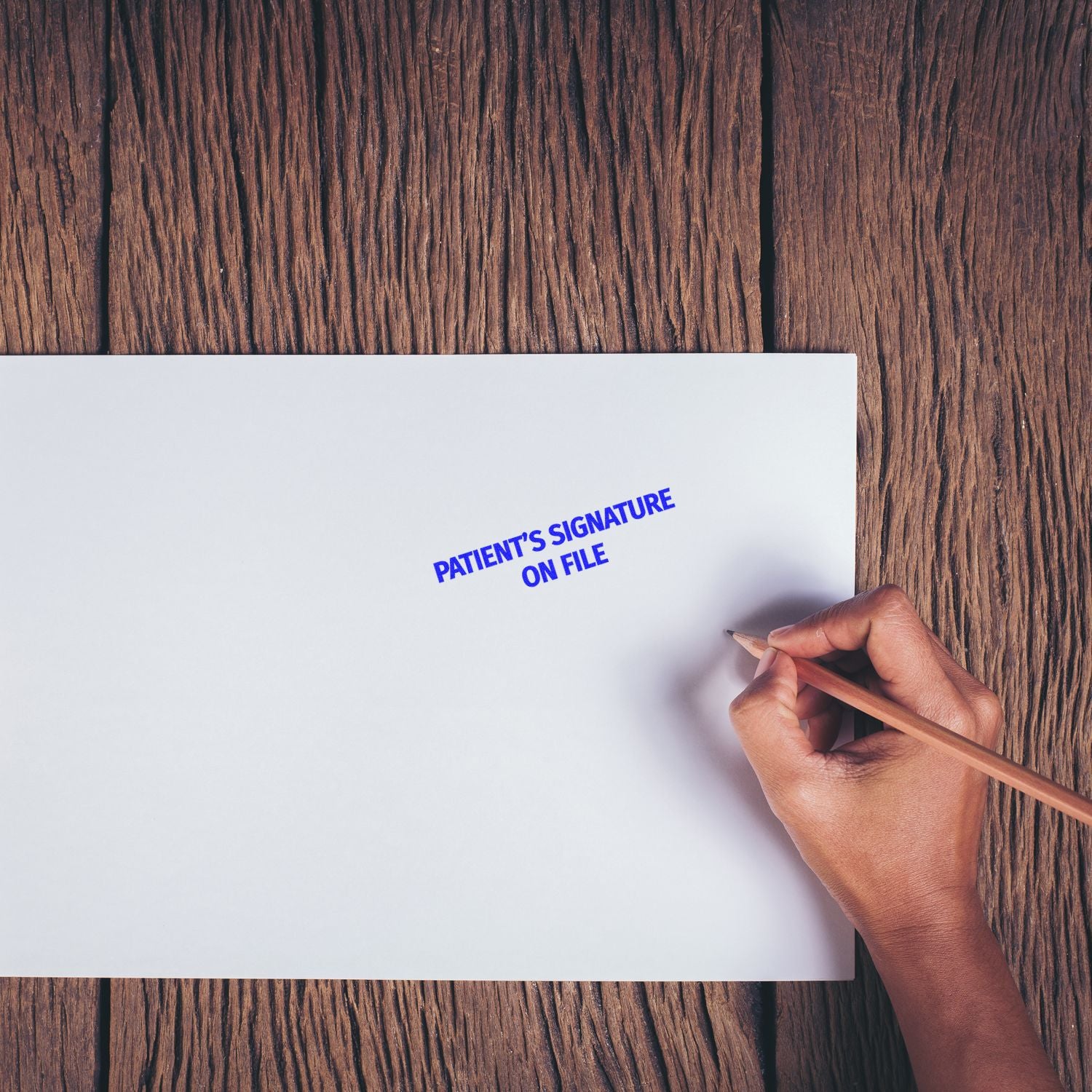 A hand holding a pencil next to a Large Pre-Inked Patients Signature on File Stamp imprint on white paper, placed on a wooden surface.