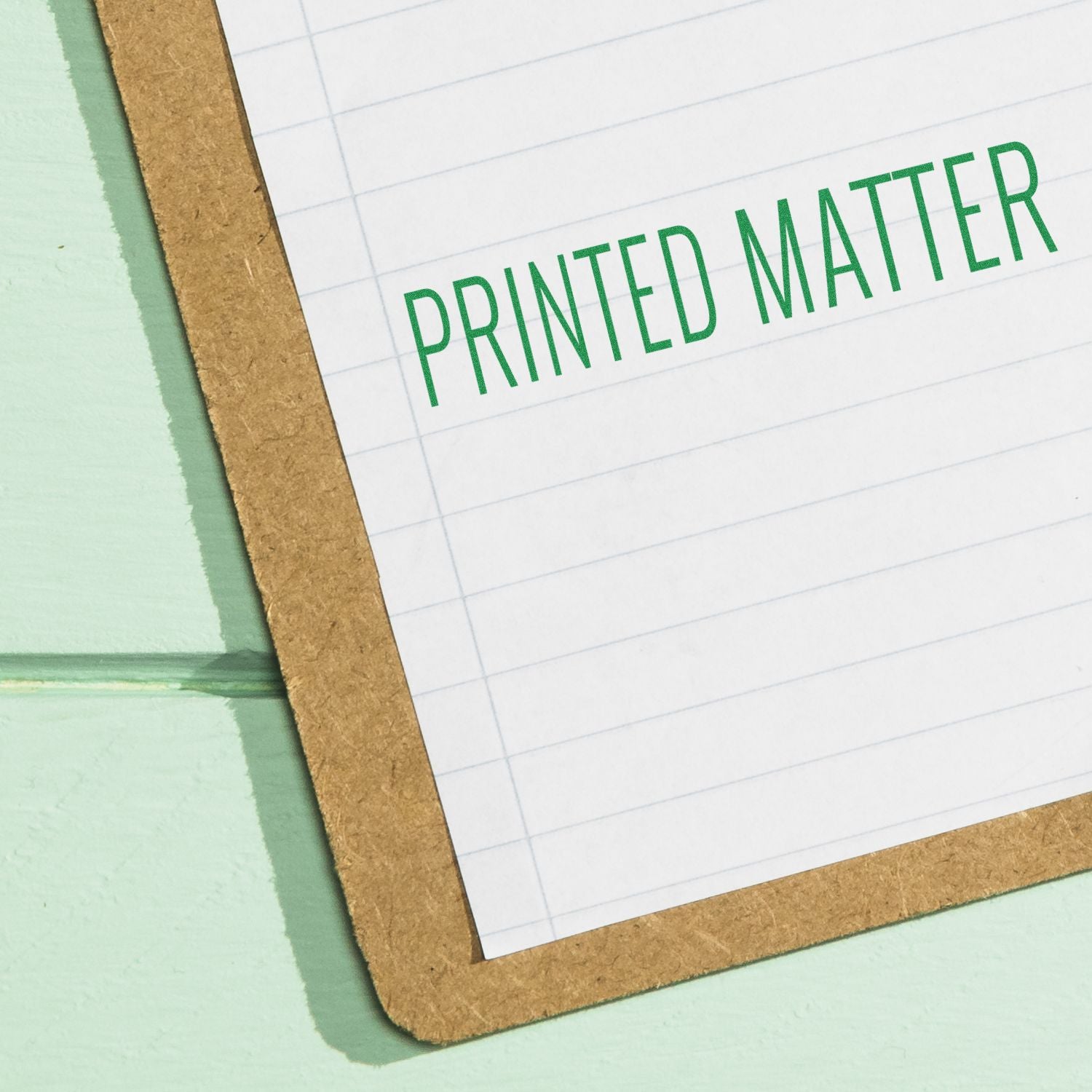 Large Pre-Inked Printed Matter Stamp imprint in green ink on a lined paper clipped to a brown clipboard, placed on a light green surface.