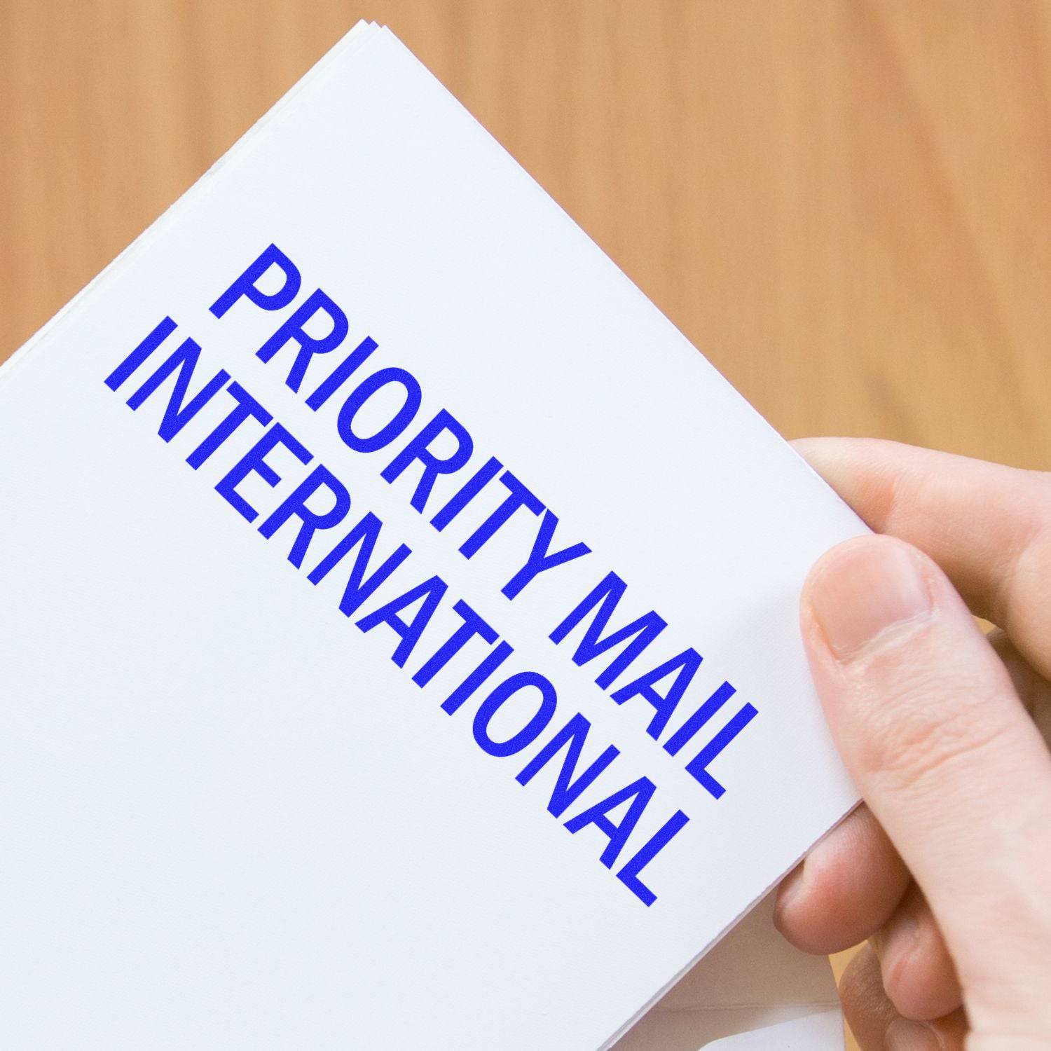 A hand holding a Large Pre-Inked Priority Mail International Stamp with blue text on a white background.