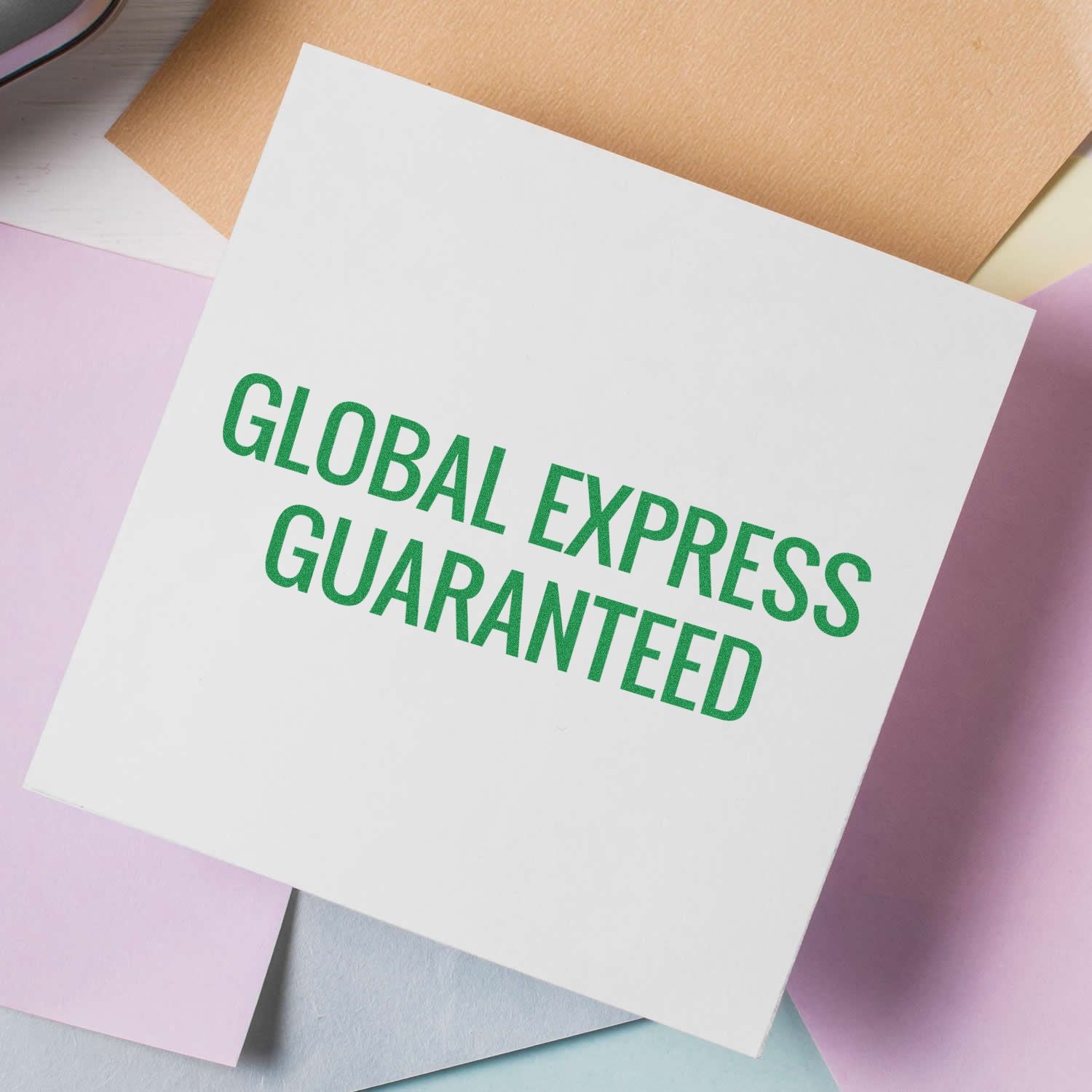 Large Pre-Inked Global Express Guaranteed Stamp on a white square paper, surrounded by pastel-colored papers on a desk.