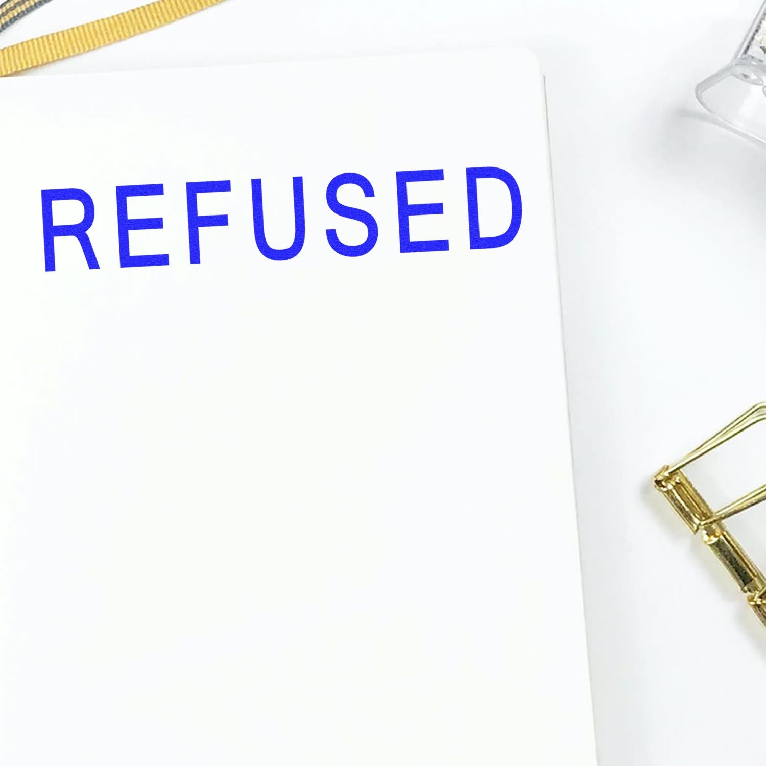 Large Pre-Inked Refused Stamp in blue ink on white paper, with a gold paperclip and a clear plastic item in the background.