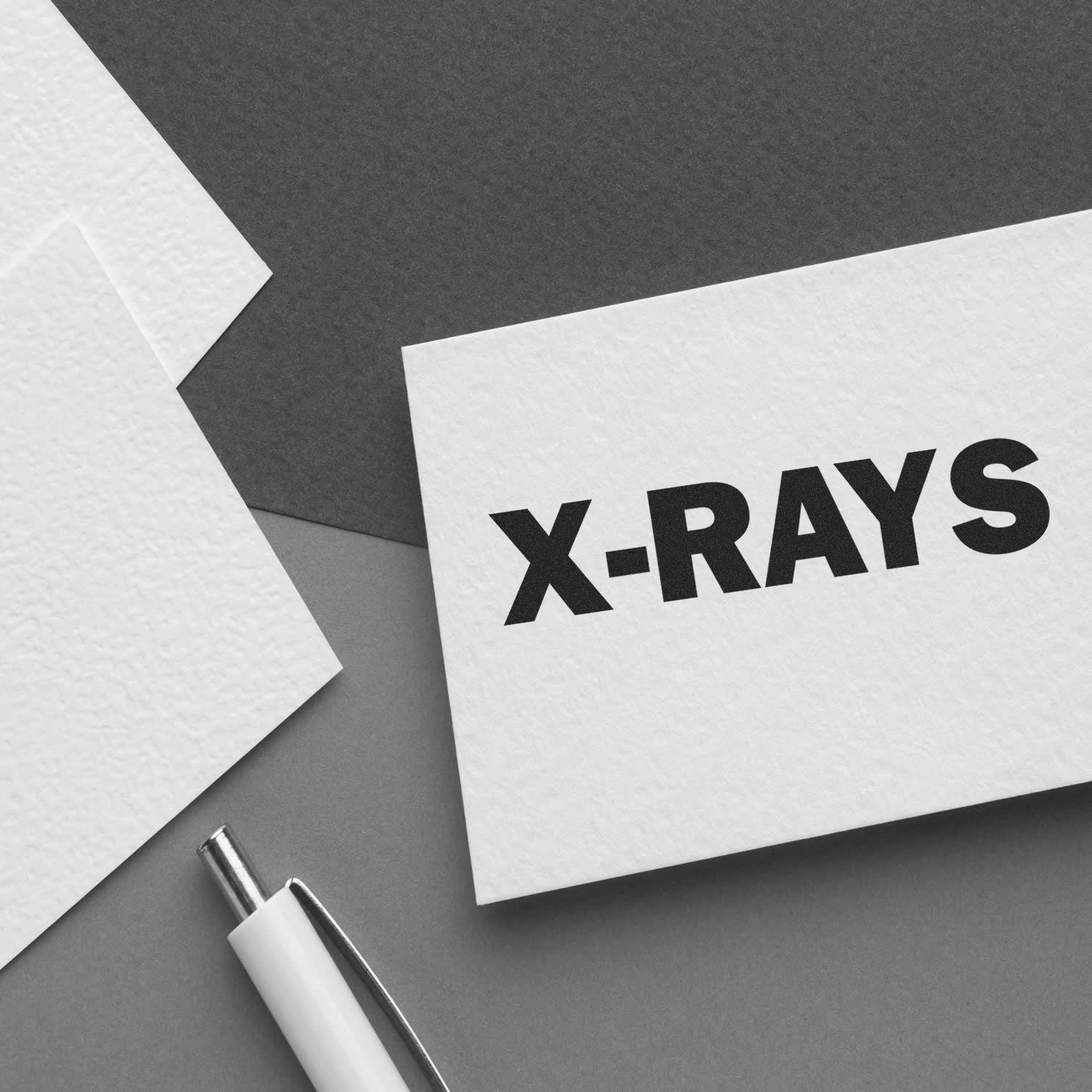 Large Pre-Inked X-Rays Stamp on a white card, placed on a gray surface with a pen and additional white cards nearby.