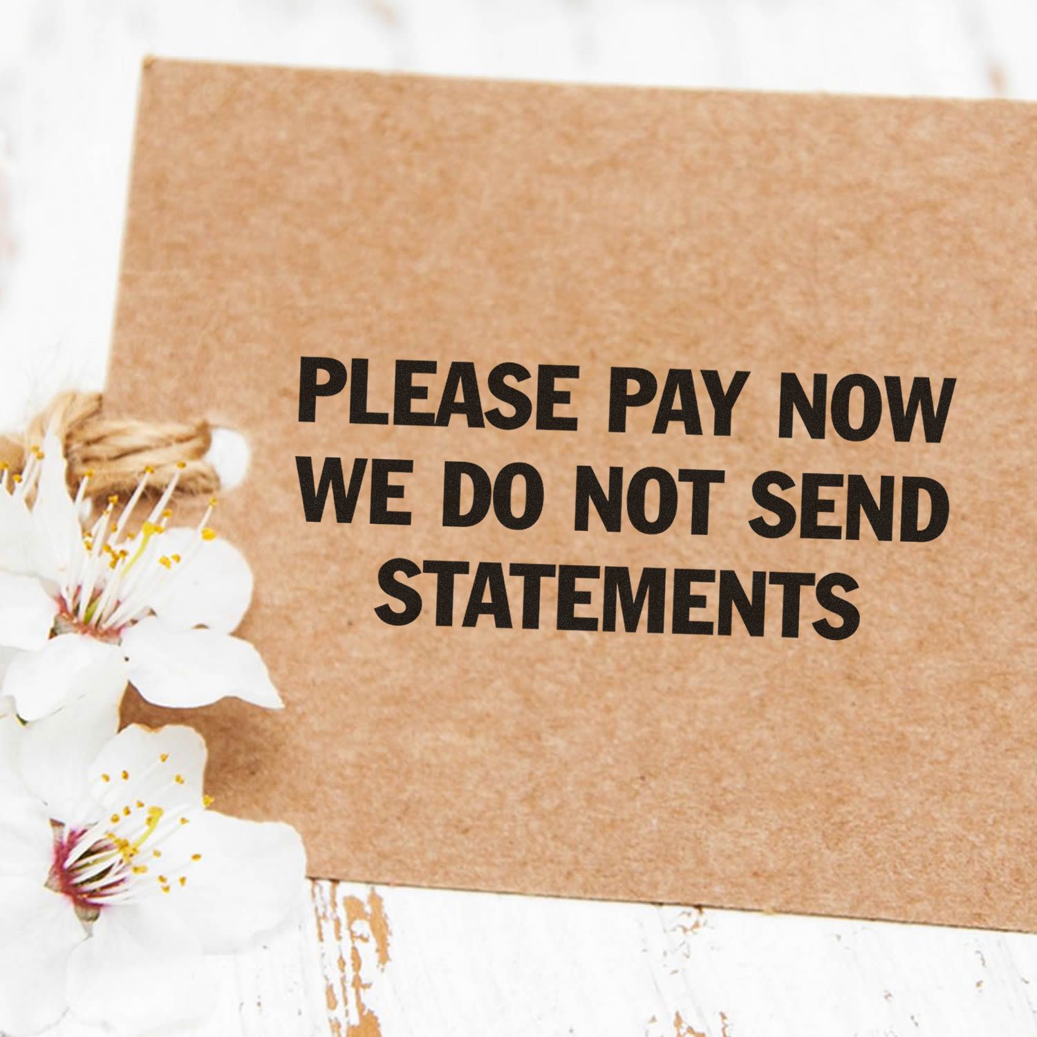 Large Pre-Inked Please Pay Now No Statements Stamp on a brown card with white flowers on a rustic white wooden surface.