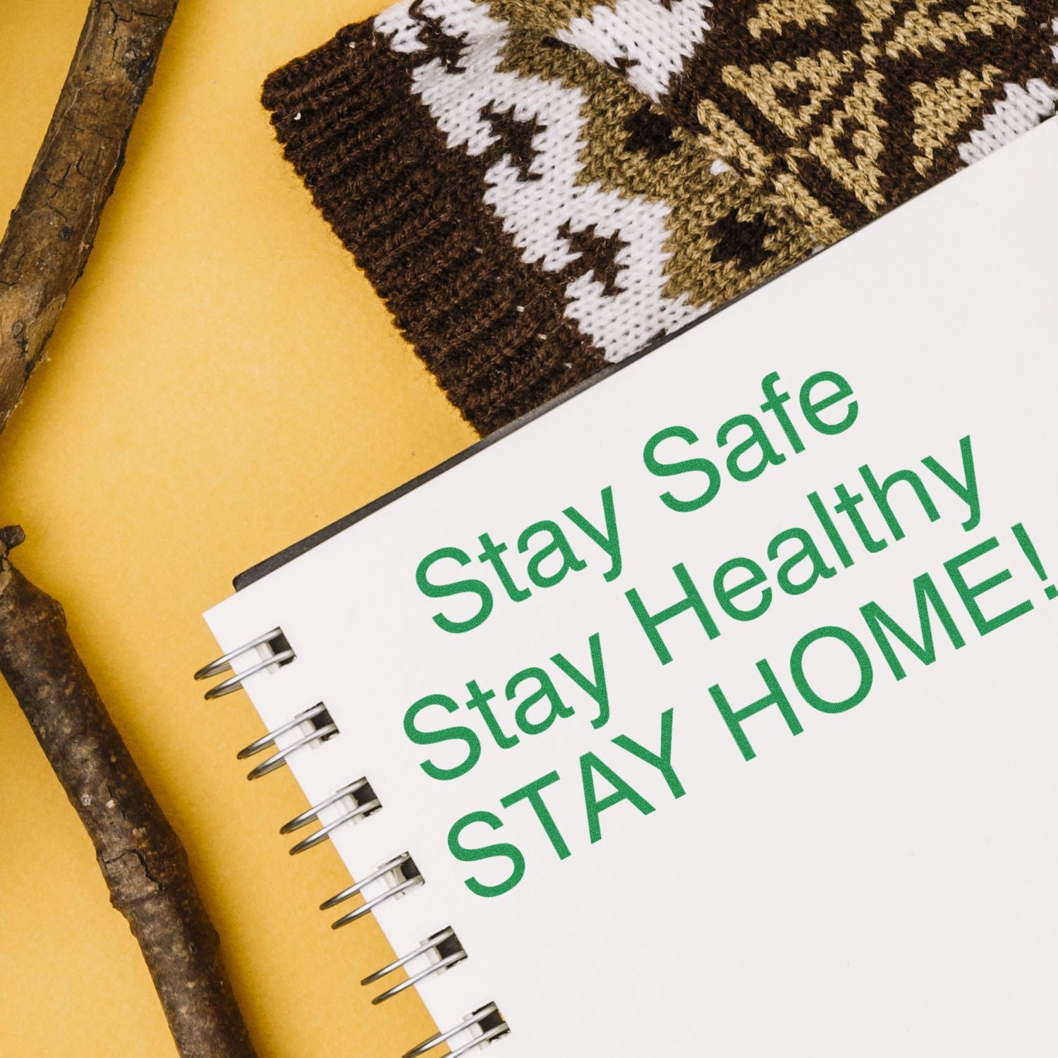 Notebook with Stay Safe Stay Healthy STAY HOME! text, next to a Large Pre-Inked Stay Safe Stay Healthy Stamp, a knitted item, and twigs.