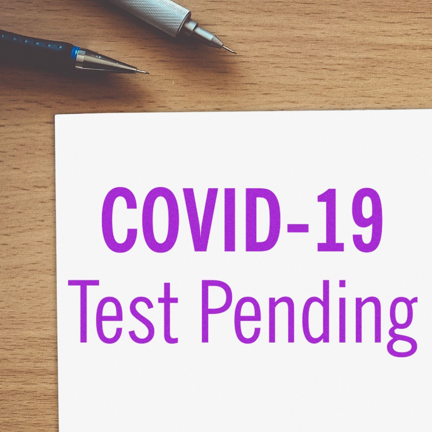 Large Pre-Inked Covid-19 Test Pending Stamp in purple ink on white paper, with a pen and pencil on a wooden desk background.