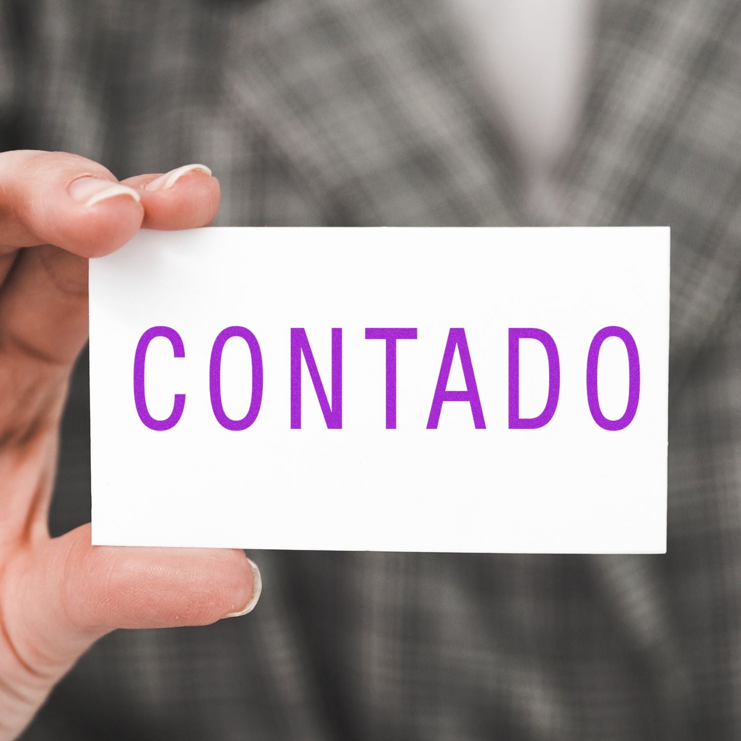 A hand holding a white card with the word CONTADO in purple letters, showcasing the Large Self Inking Contado Stamp. The background features a blurred plaid pattern.