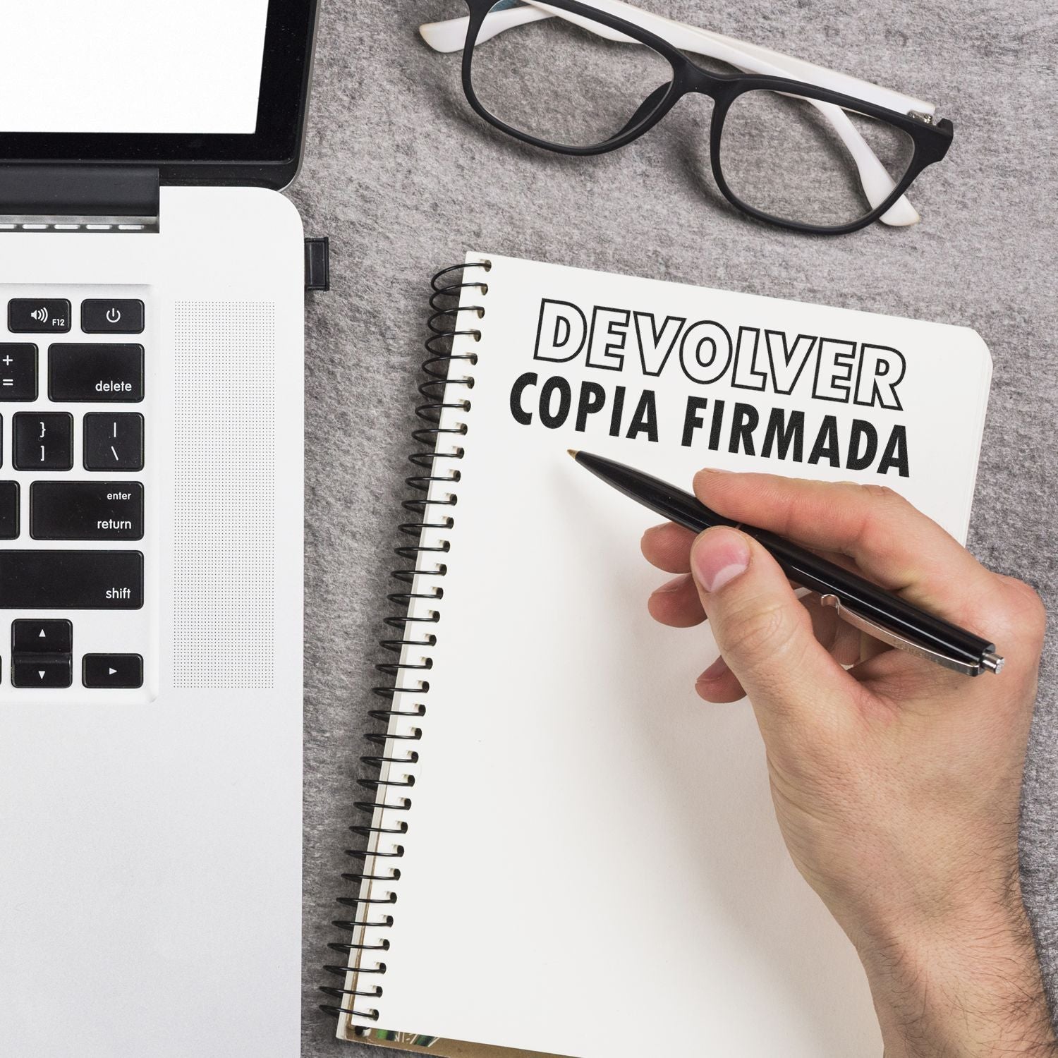 A hand holding a pen near a notebook with DEVOLVER COPIA FIRMADA text, next to a laptop and glasses. Large Self Inking Devolver Copia Stamp.