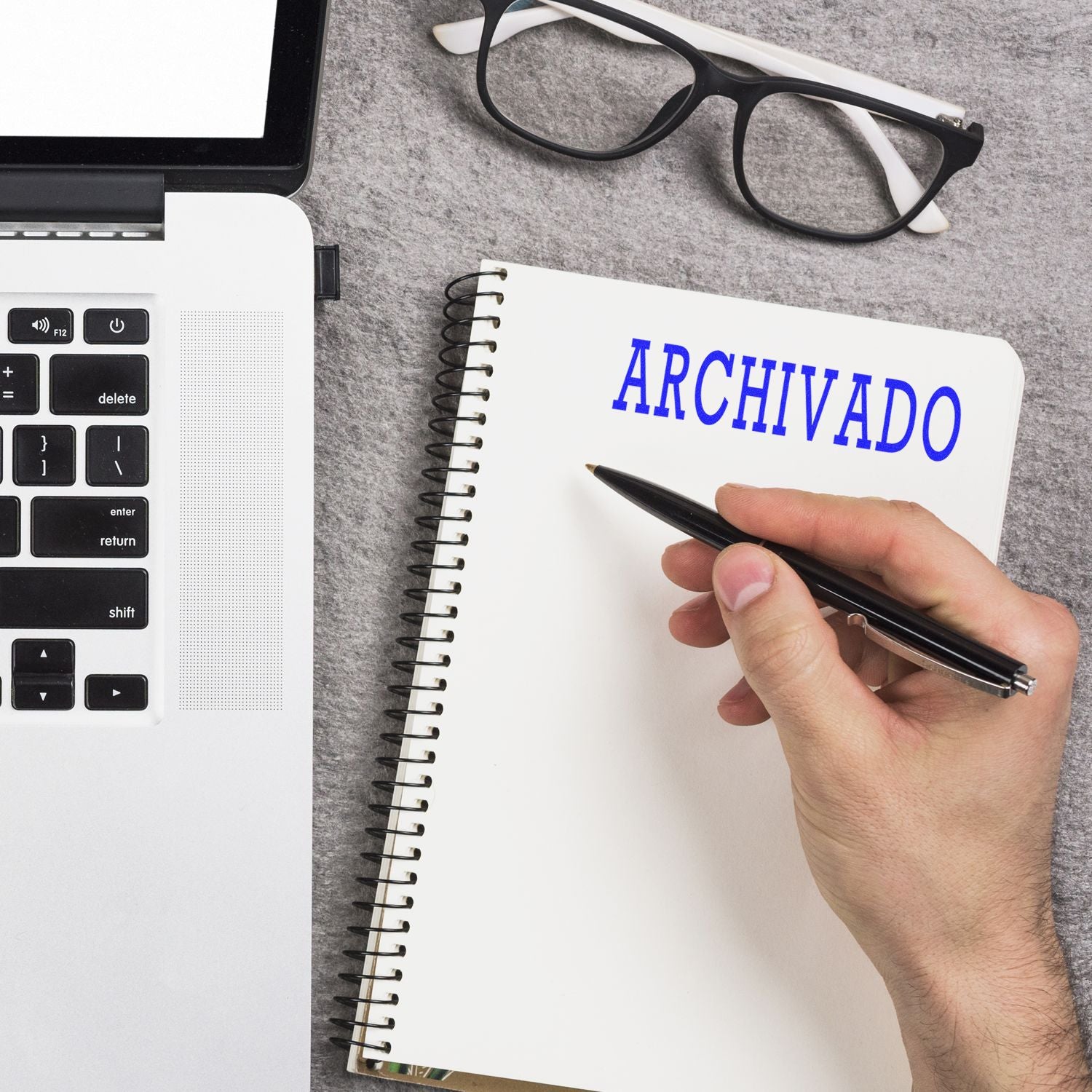 A hand writing in a notebook labeled ARCHIVADO next to a laptop, glasses, and a Large Self Inking Archivado Stamp on a gray surface.