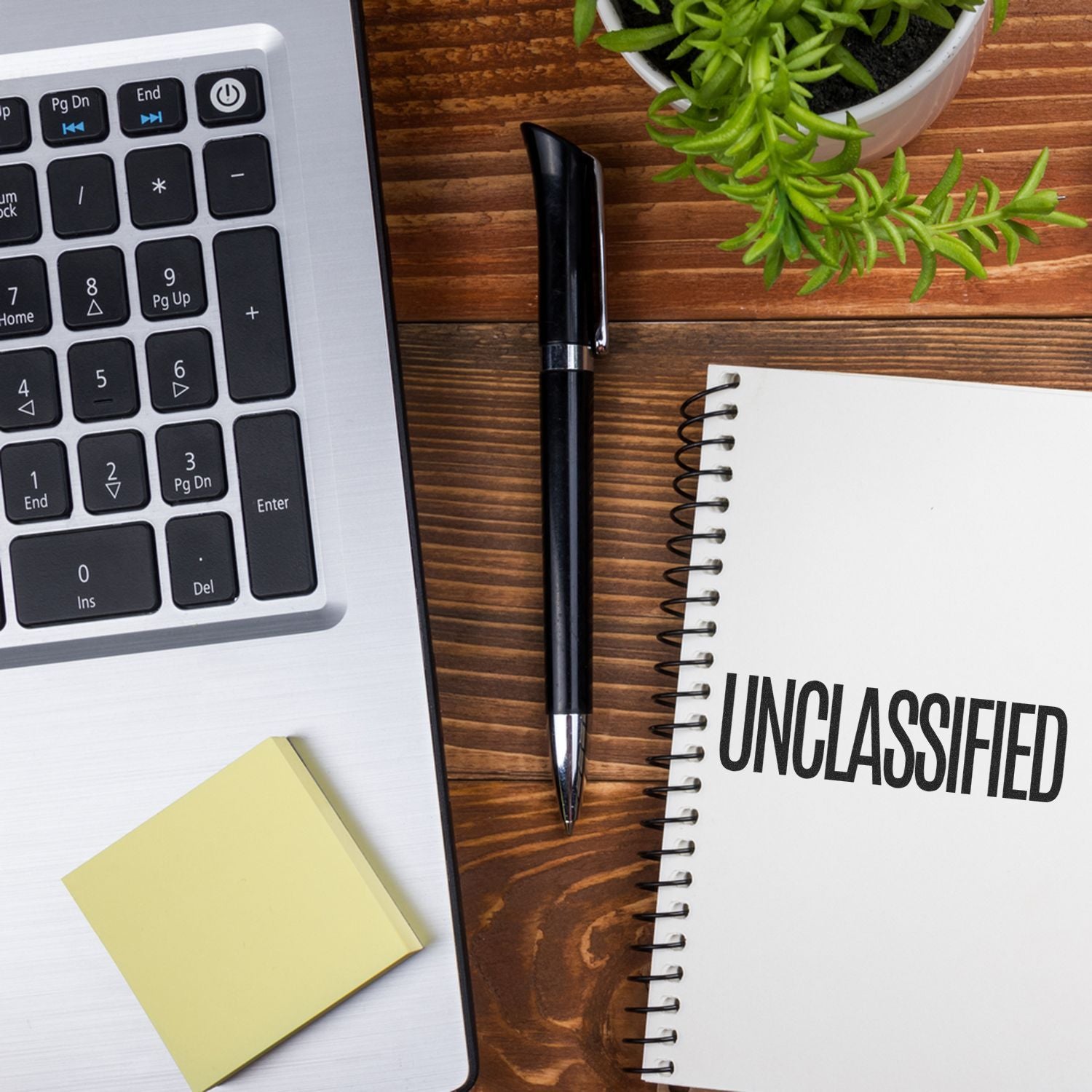 A desk with a laptop, sticky notes, pen, plant, and a notebook stamped with UNCLASSIFIED using the Large Pre-Inked Unclassified Stamp.